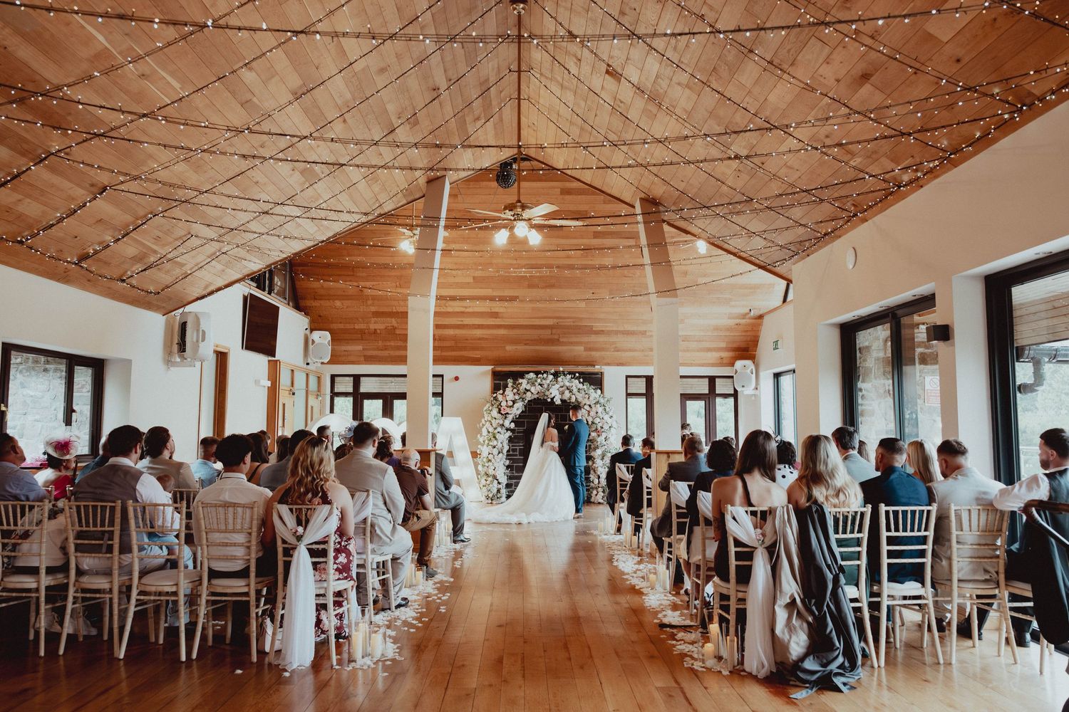 Elegant indoor wedding ceremony in rustic venue with fairy lights, floral arch, and seated guests.