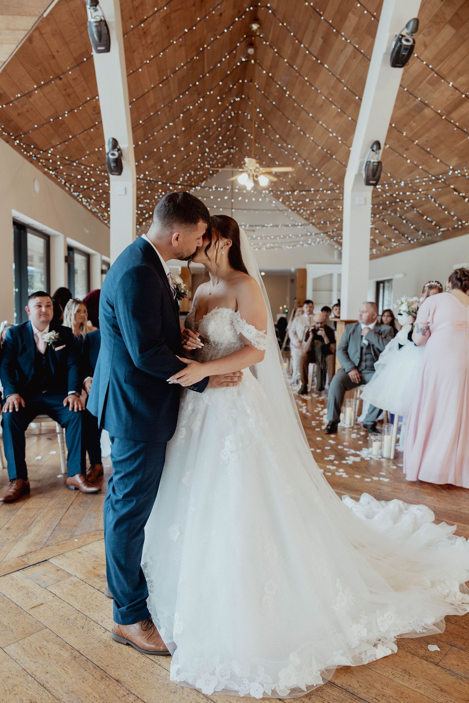 Newlyweds share first kiss on petal-strewn aisle under twinkling fairy lights in rustic wedding venue in Canada lodge south Wales.