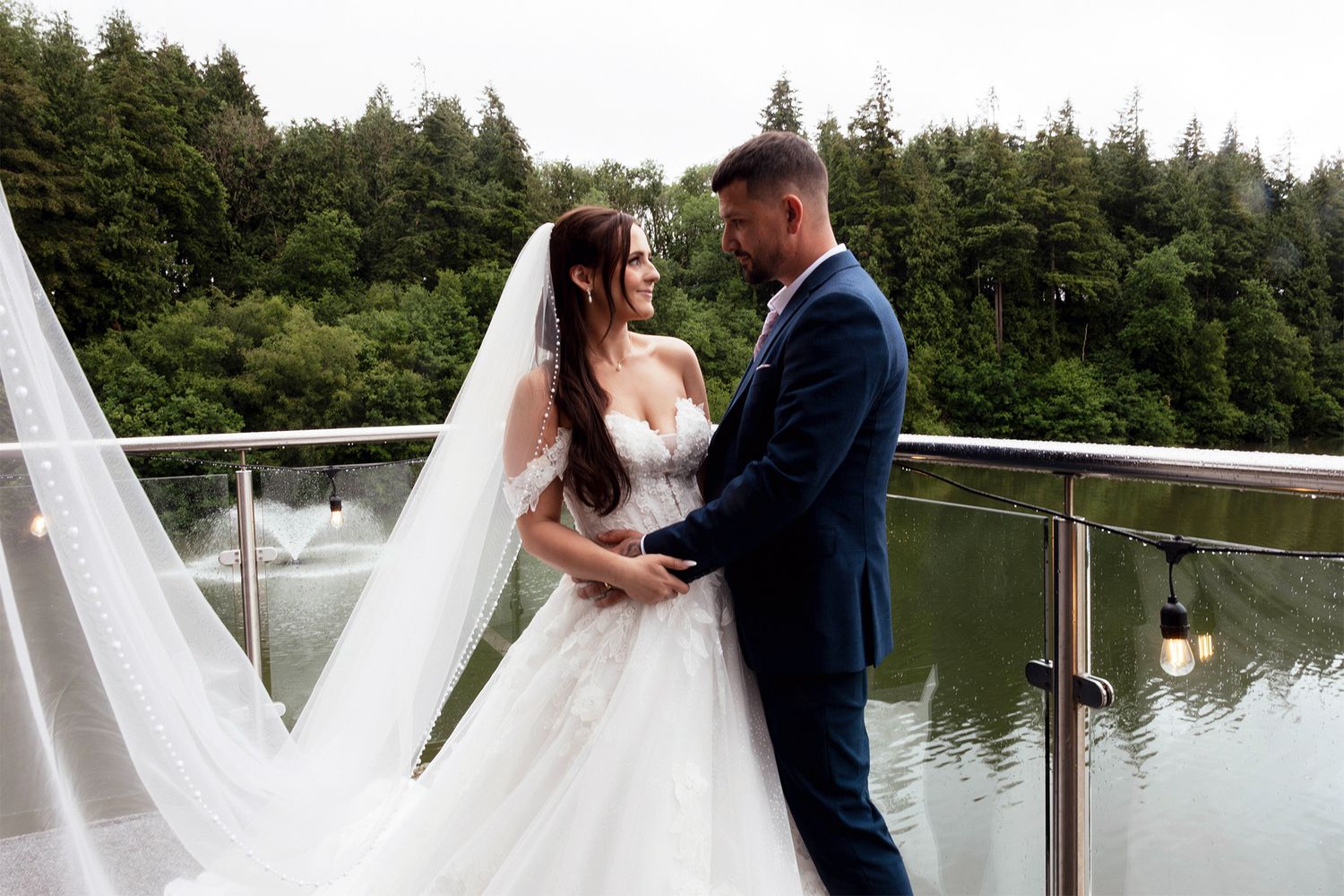 Bride in lace gown with long veil and groom in navy suit share romantic moment on lakeside balcony.