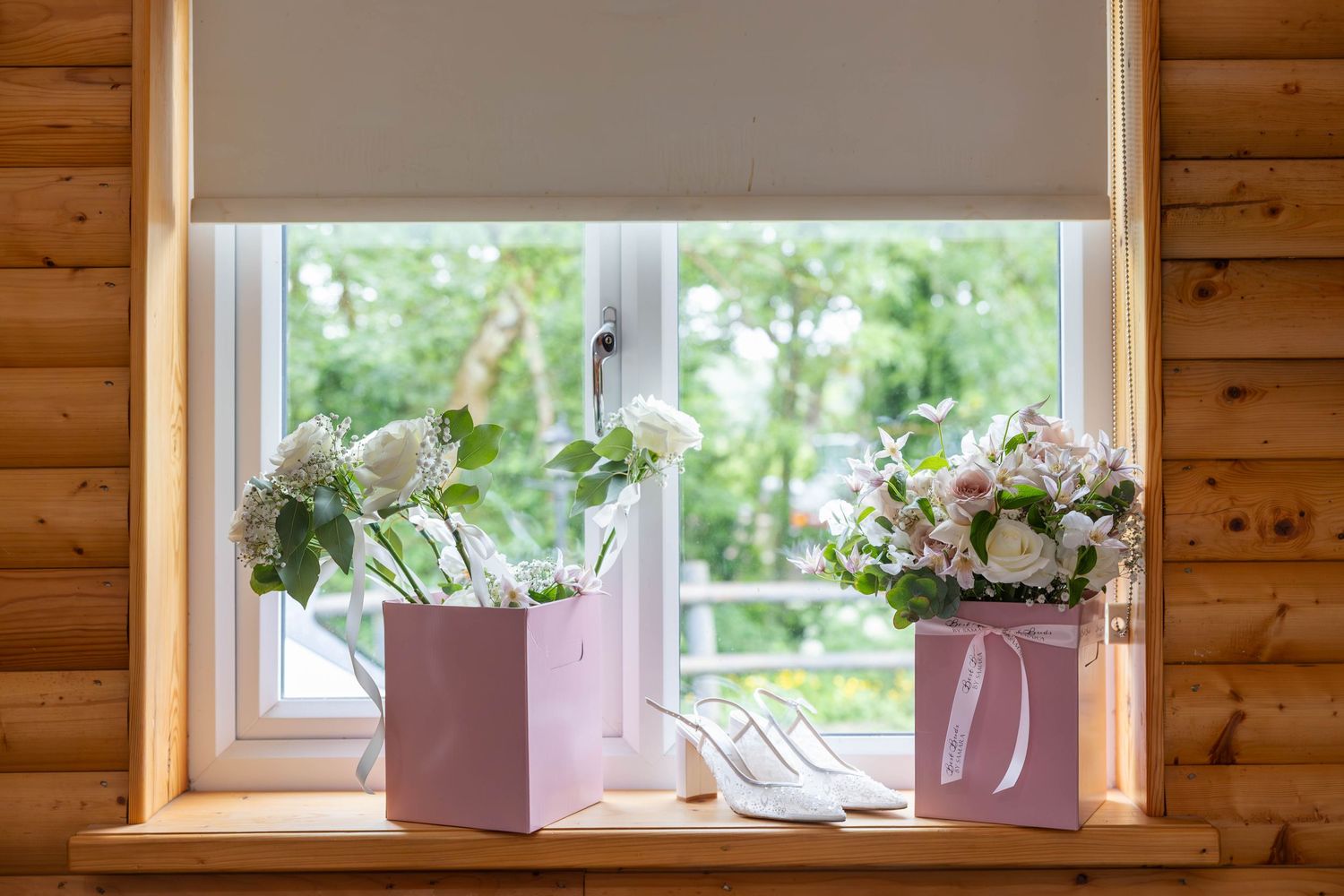 Bridal accessories displayed on windowsill with white heels and two pink gift bags with floral bouquets.