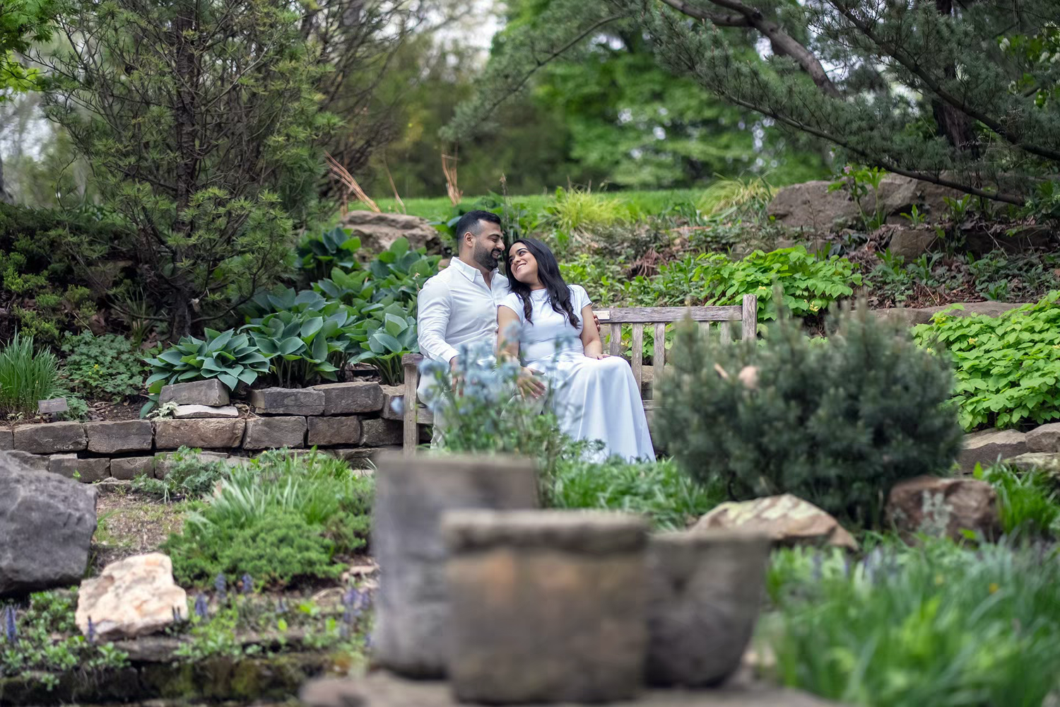 Couple in white attire sitting on a bench surrounded by lush greenery in Inniswood Metro Gardens Engagement Photos