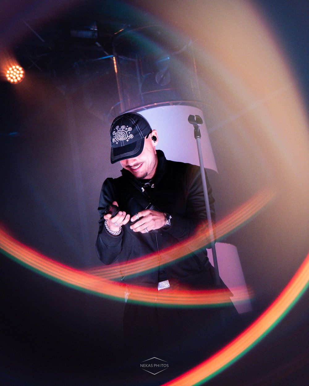 DJ in black cap and jacket looks at phone under colorful light trails at a nightclub event.