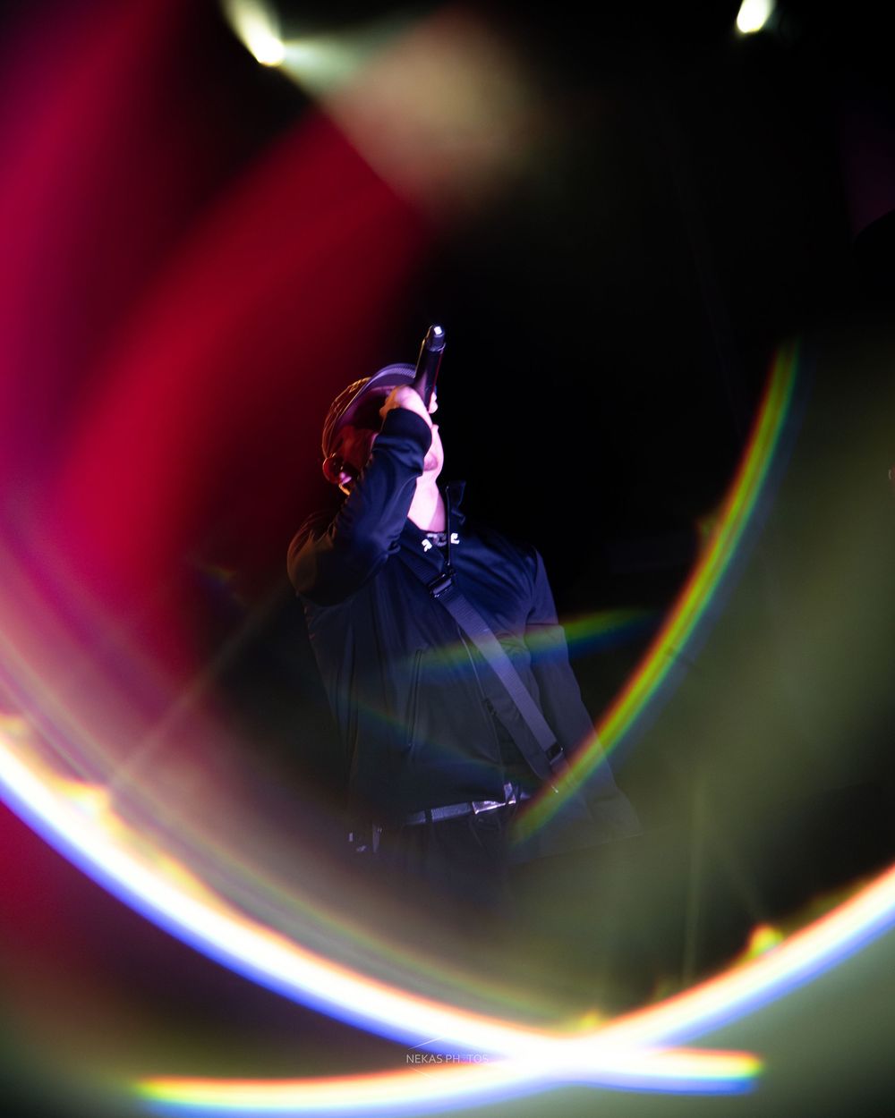 Performer singing on stage surrounded by vibrant colorful lens flares and rainbow light effects in a dark concert venue.