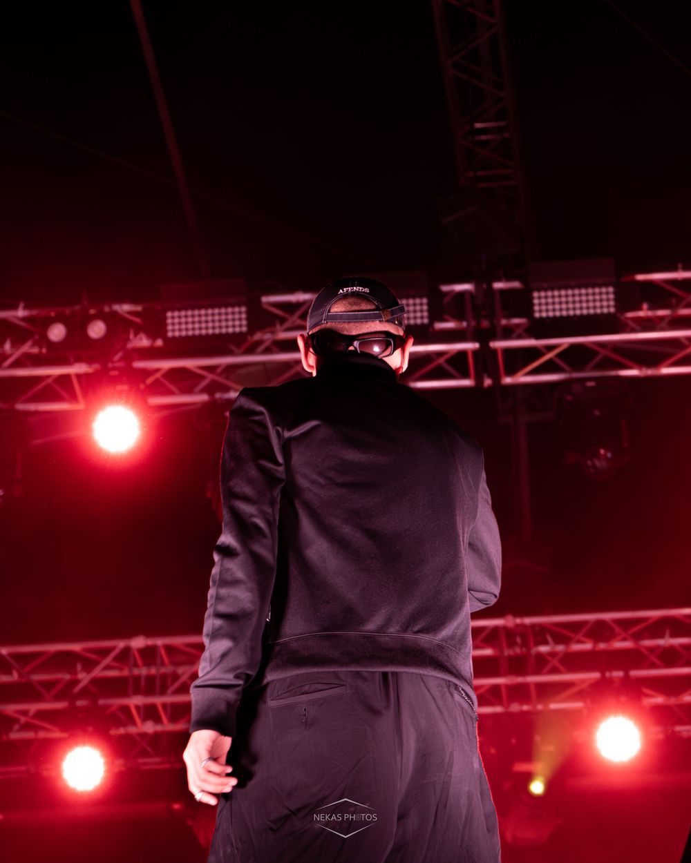 Performer in all-black outfit and mask stands on stage under dramatic red lighting at an outdoor concert event.