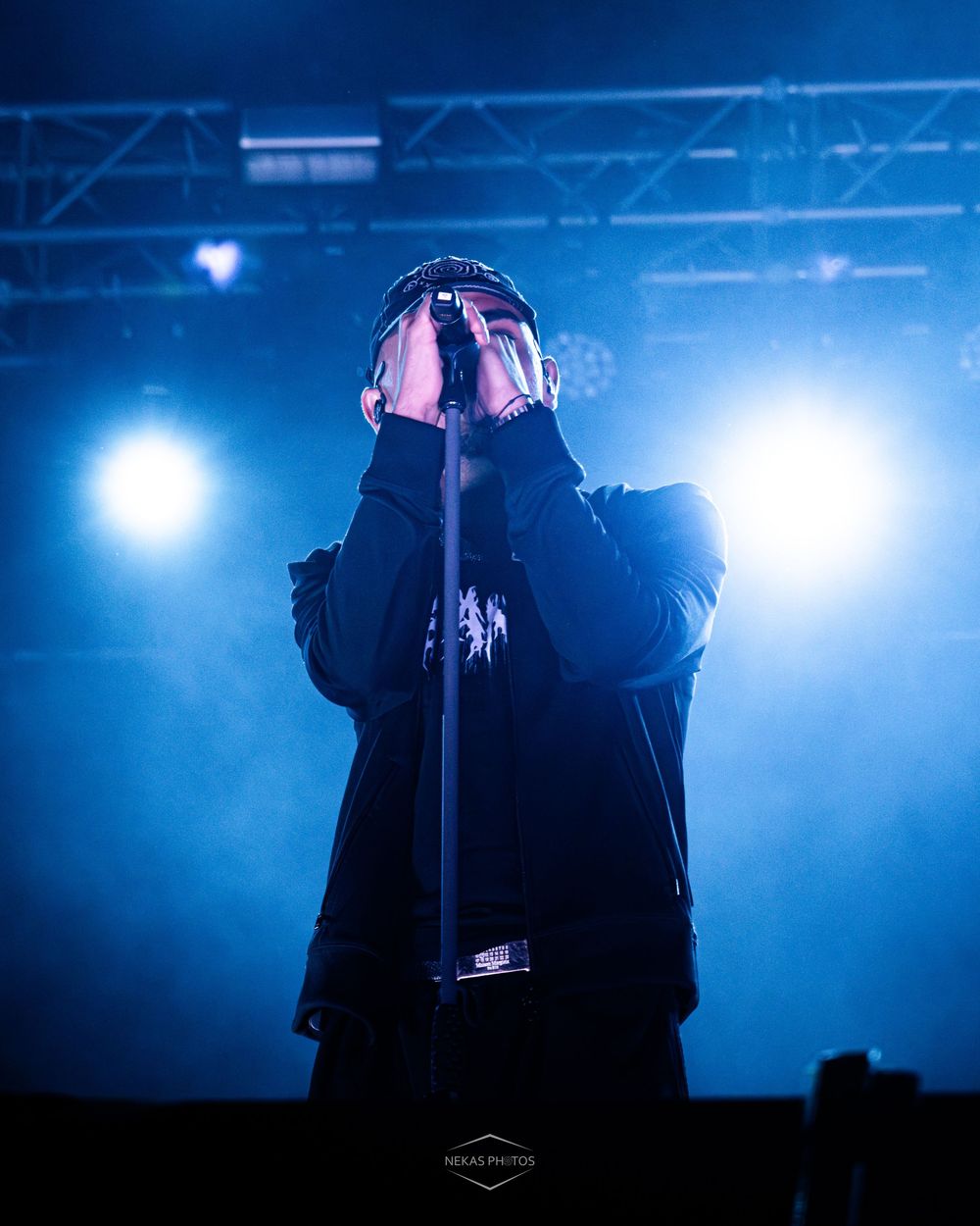 A dramatic concert performer holds a microphone stand on a fog-filled stage bathed in deep blue and white spotlights.