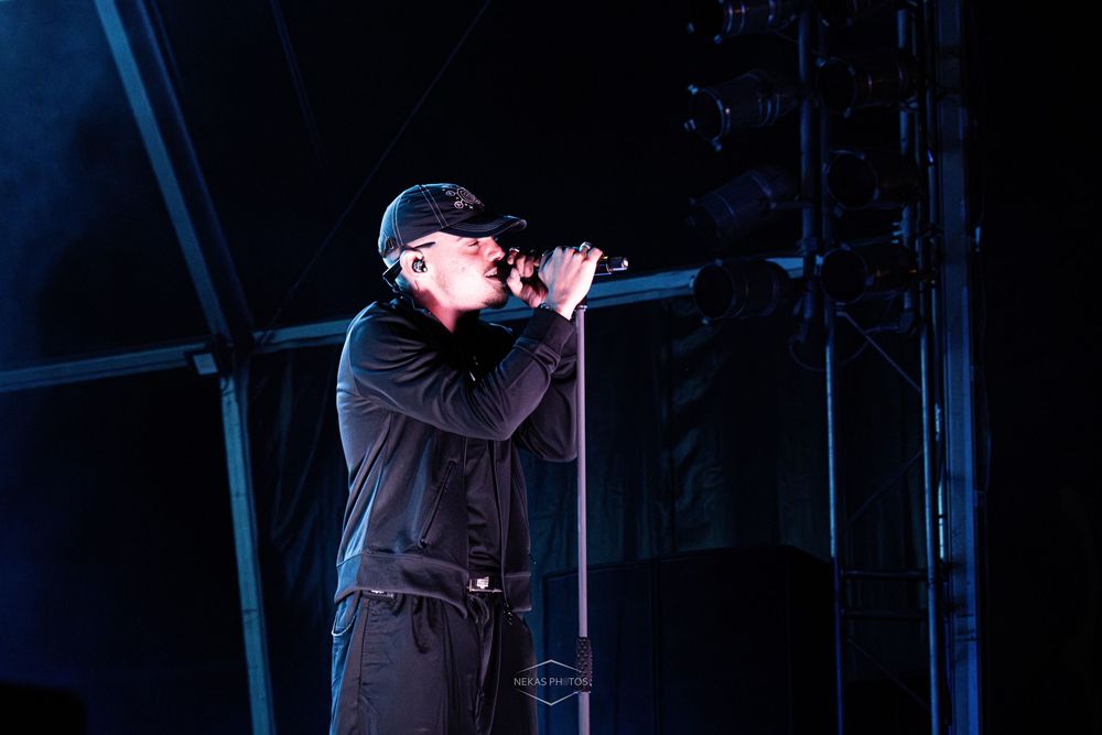 Male singer in black outfit and cap performing on stage with microphone under blue stage lighting.