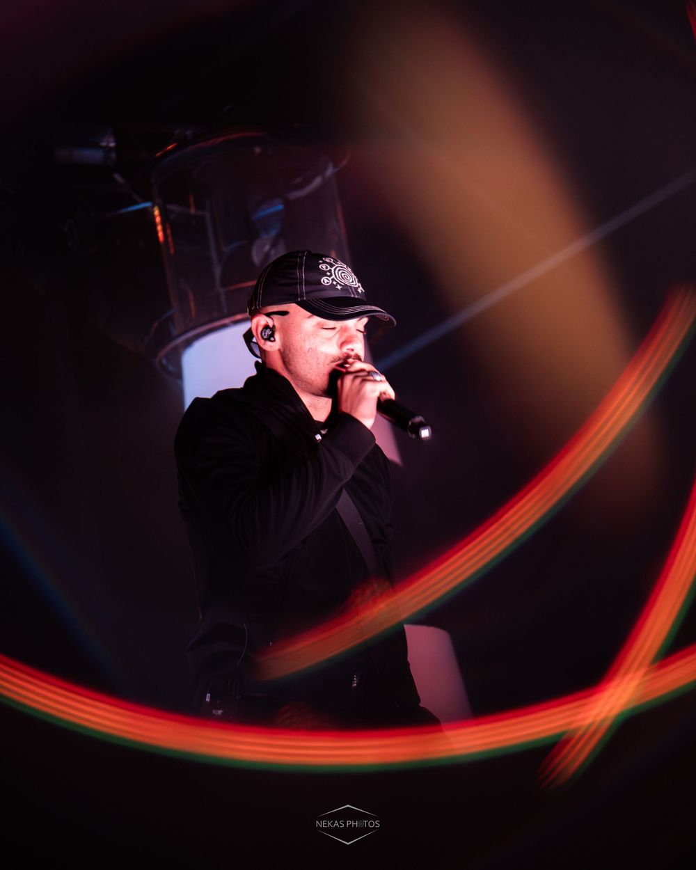 Male singer performing on stage with microphone, wearing black cap, surrounded by colorful light streaks in dark venue.