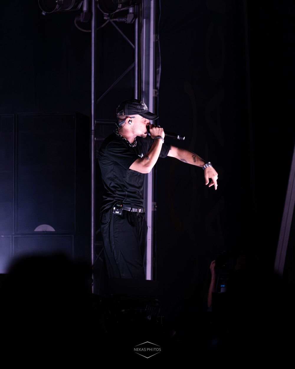 A performer in all-black outfit holds a microphone on a dimly lit stage with purple lighting during a live concert.