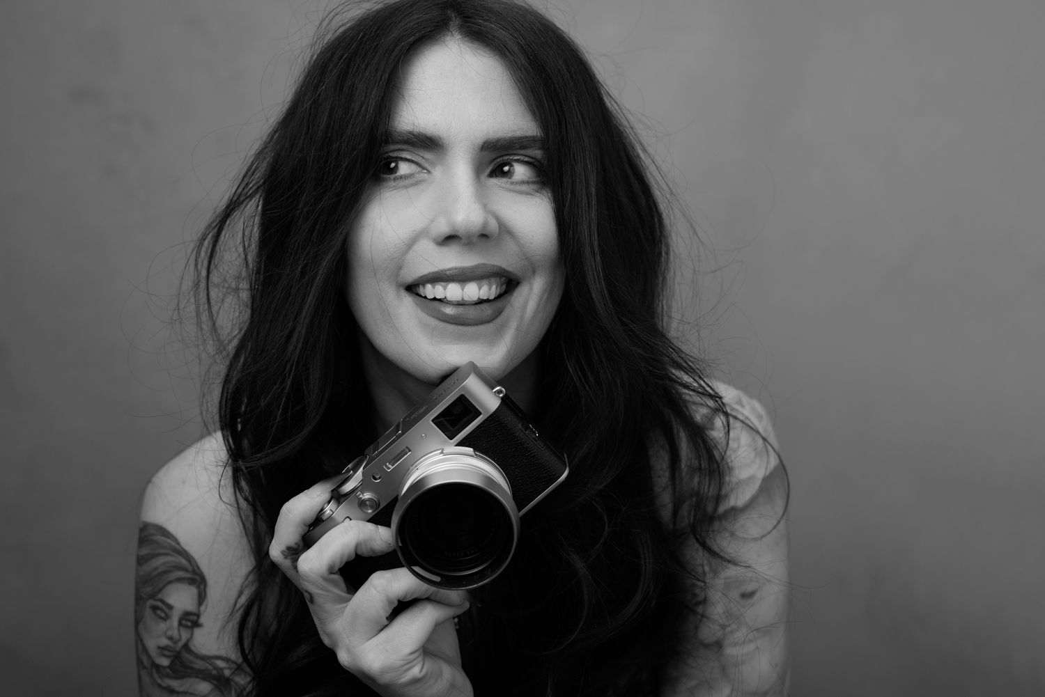 Black and white photo of a smiling woman with long dark hair holding a camera, showing tattoos on her shoulder.