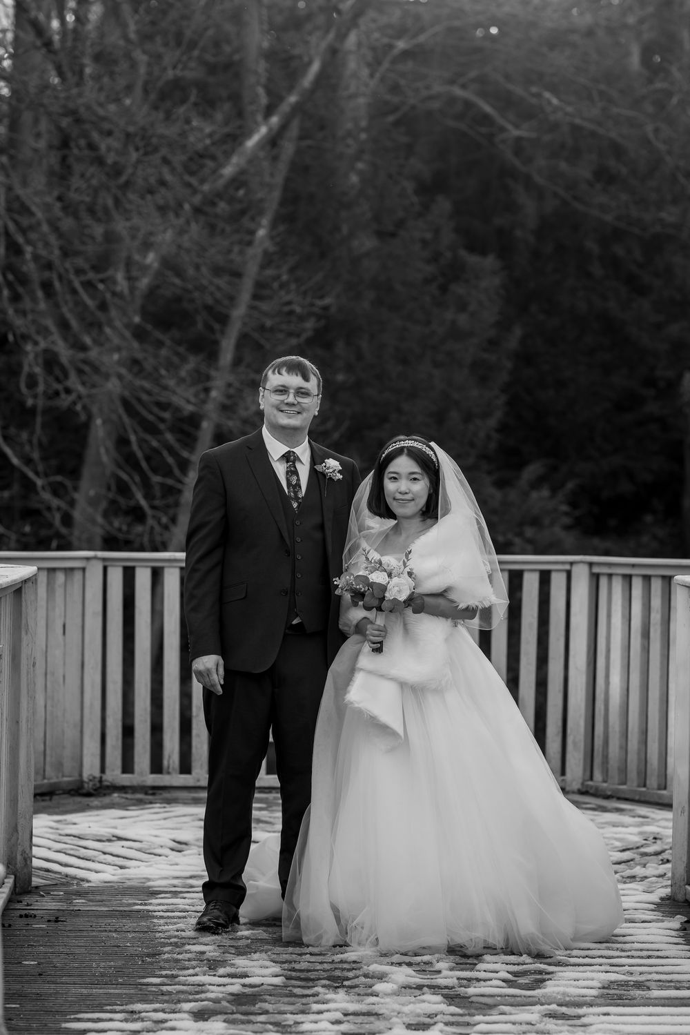 Black and white wedding photo of bride and groom standing on wooden deck surrounded by trees.
