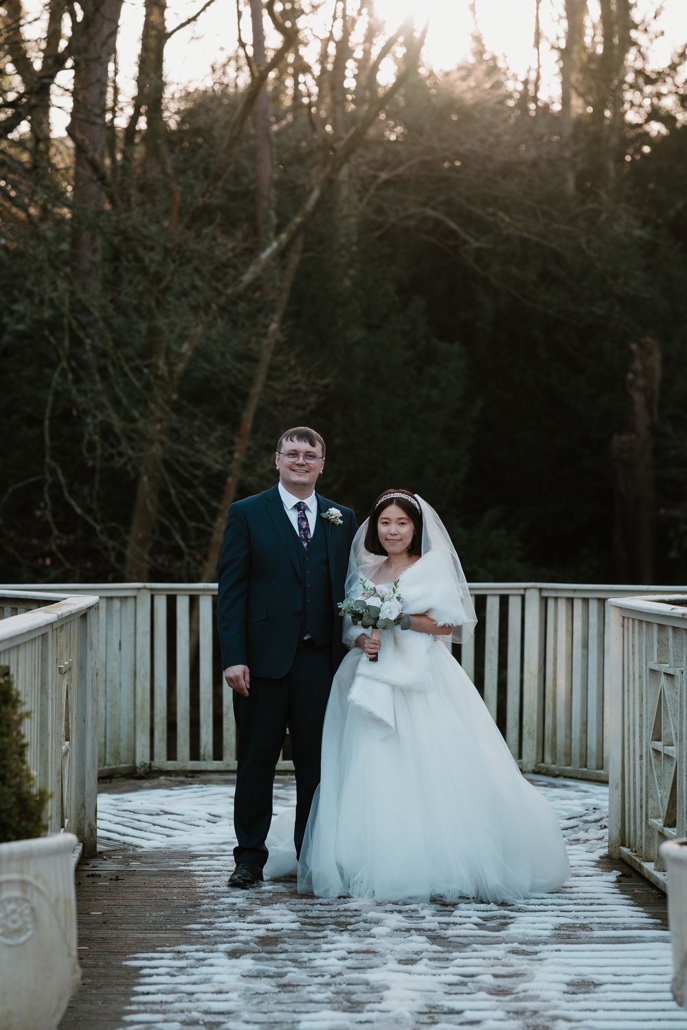 Bride and groom posing on a snowy wooden bridge in winter, surrounded by bare trees in golden hour light.