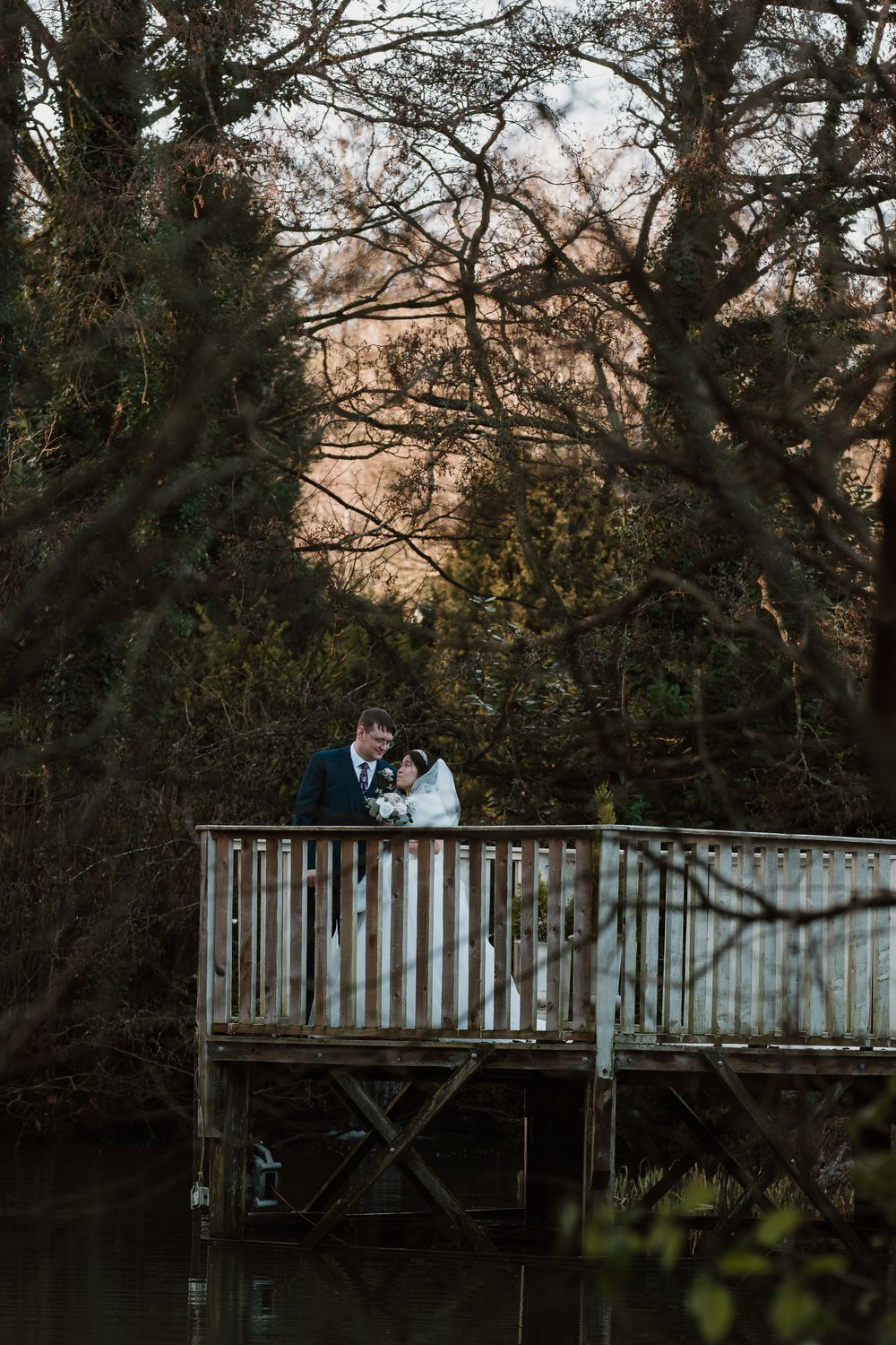 Bride and groom embracing on a wooden dock surrounded by lush trees and a serene pond at dusk.