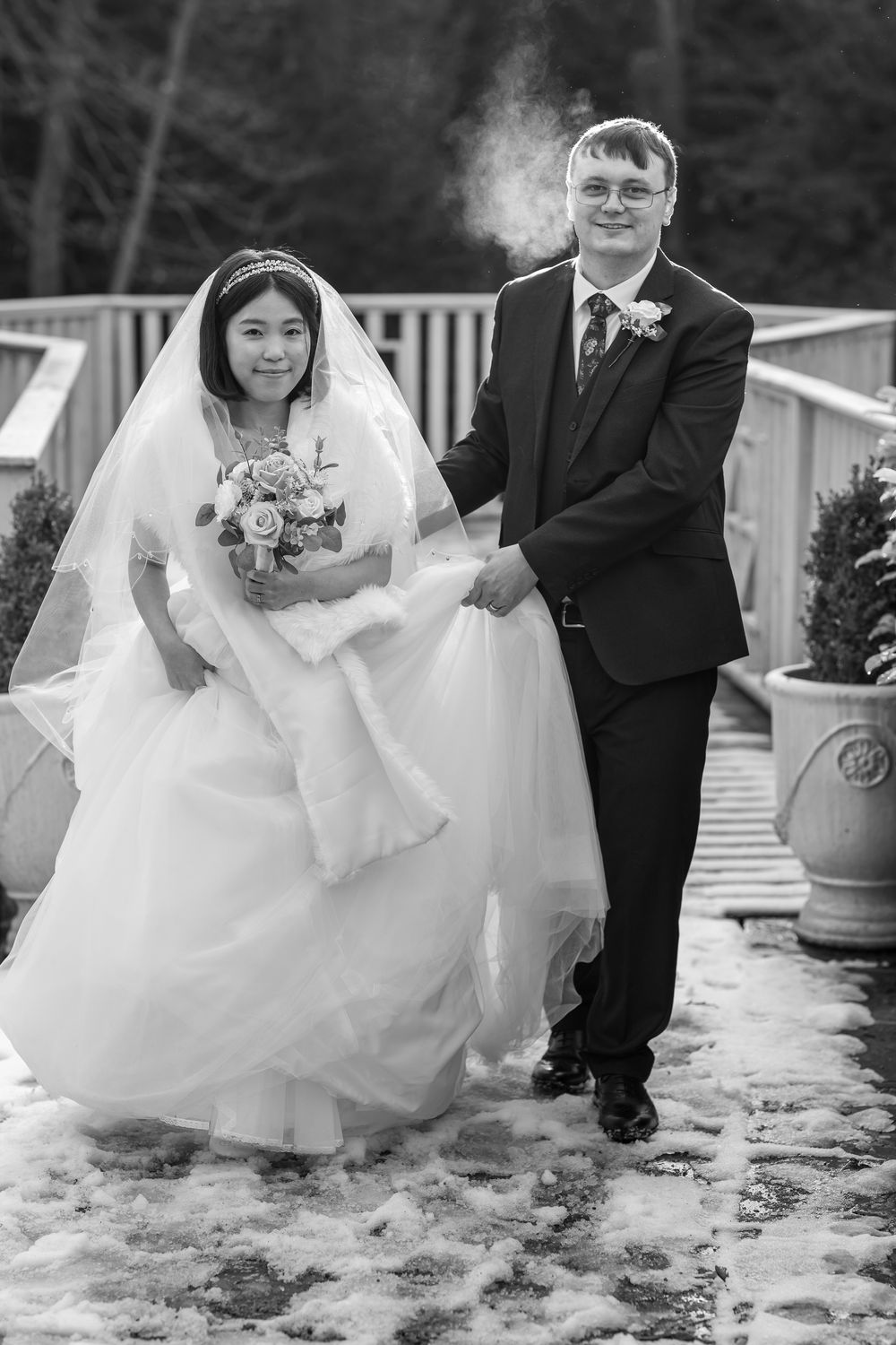 Black and white photo of a bride and groom walking outdoors in snow, bride holding bouquet in winter wedding attire.