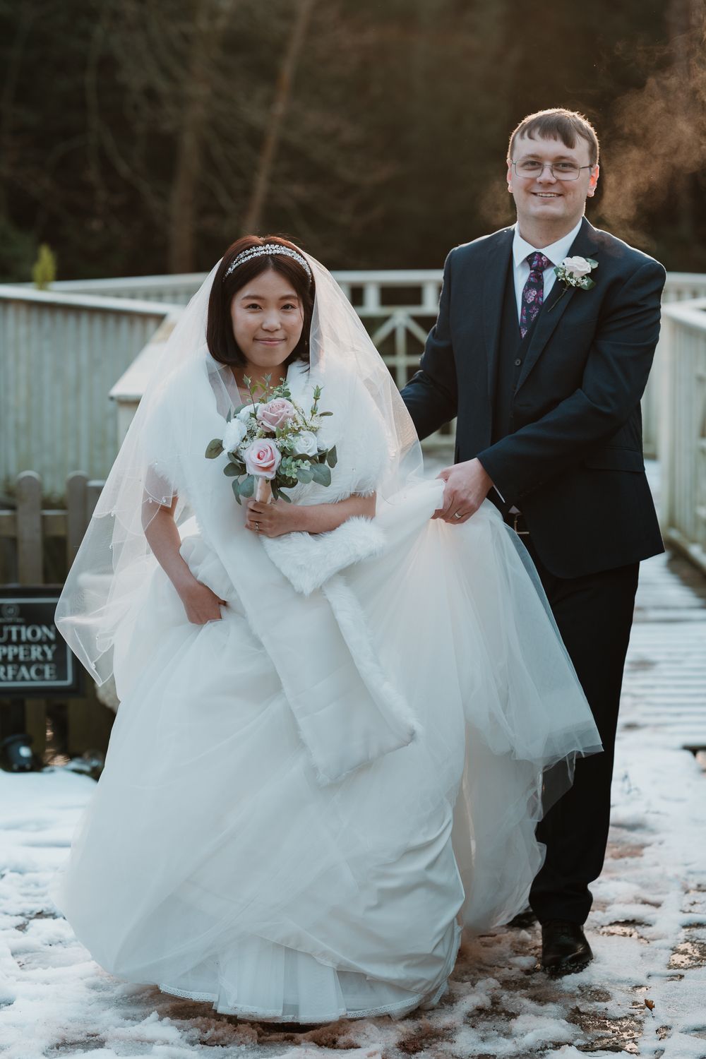 Bride in white gown holding bouquet and groom in dark suit walking on snowy bridge in winter wedding photo.