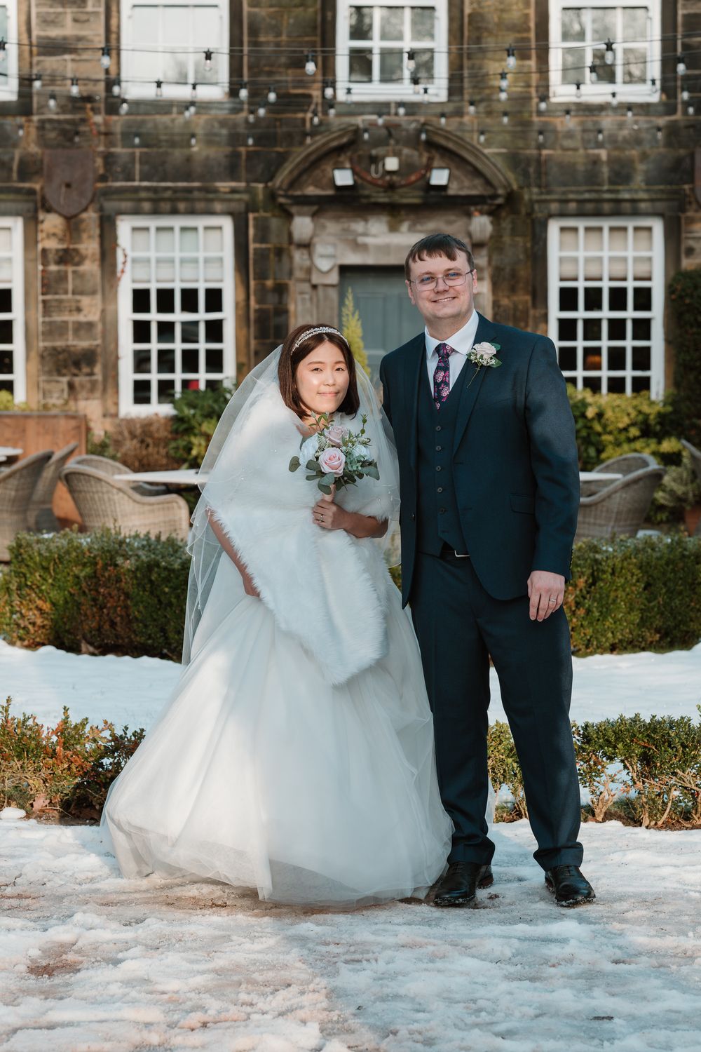 Bride in white gown with fur wrap and groom in navy suit pose together in snowy garden outside stone manor house.