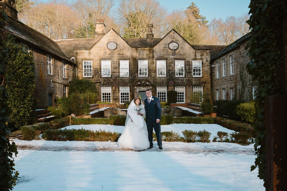 Bride and groom standing in snowy winter garden outside historic stone manor house wedding venue.