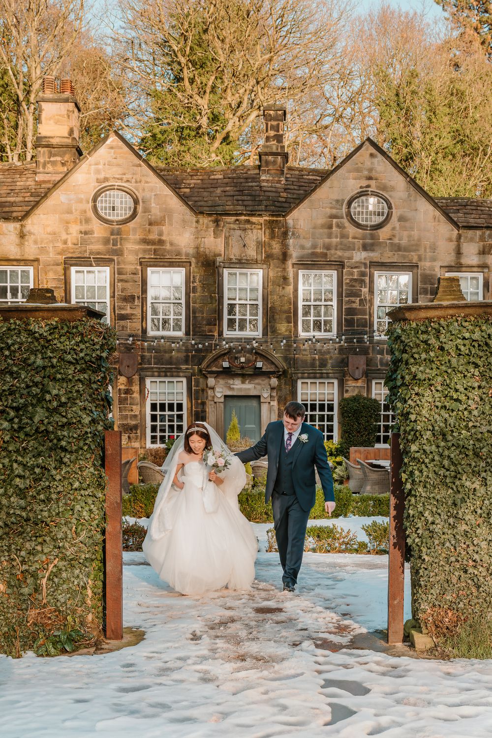 Bride and groom walking hand in hand through snowy garden gates at a historic stone manor in winter.