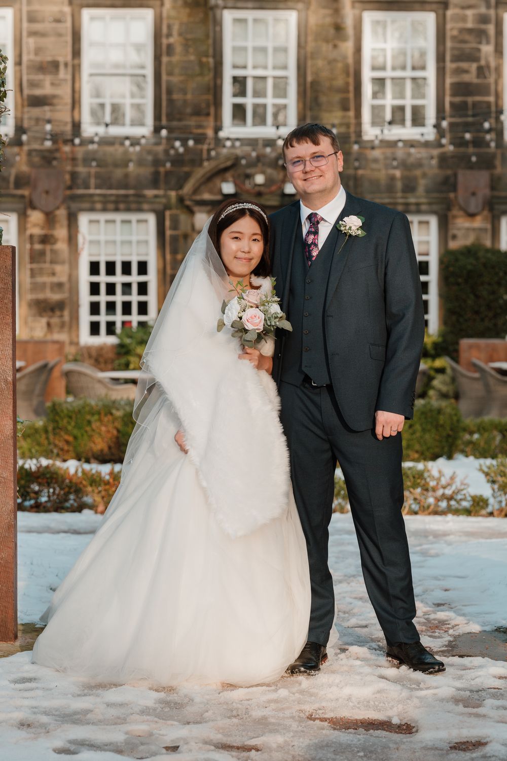 Bride and groom posing outdoors in winter snow, bride in white gown with fur wrap holding bouquet, groom in dark suit.