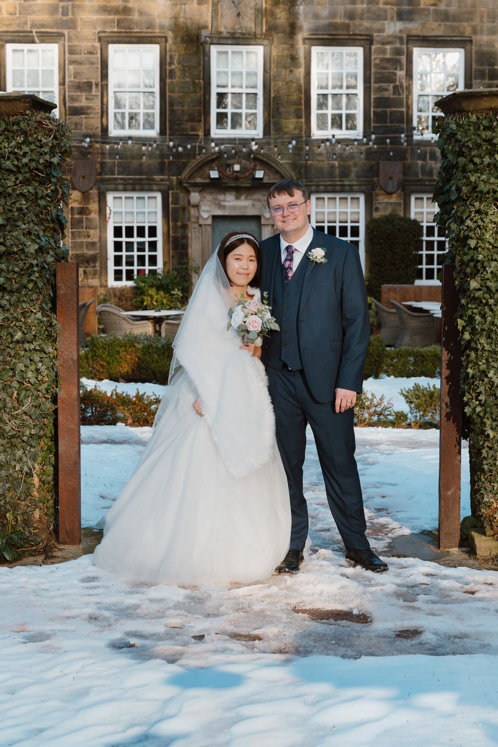 A bride and groom pose together in the snow outside a historic stone manor house in a winter wedding portrait.