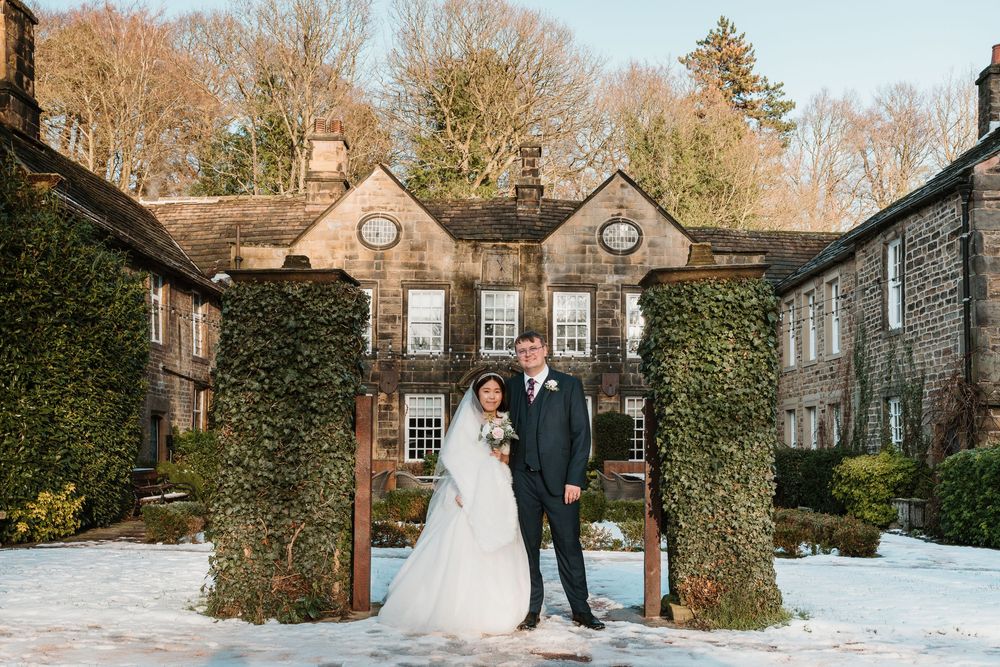 Bride and groom pose in snowy winter garden outside ivy-covered stone manor house wedding venue.