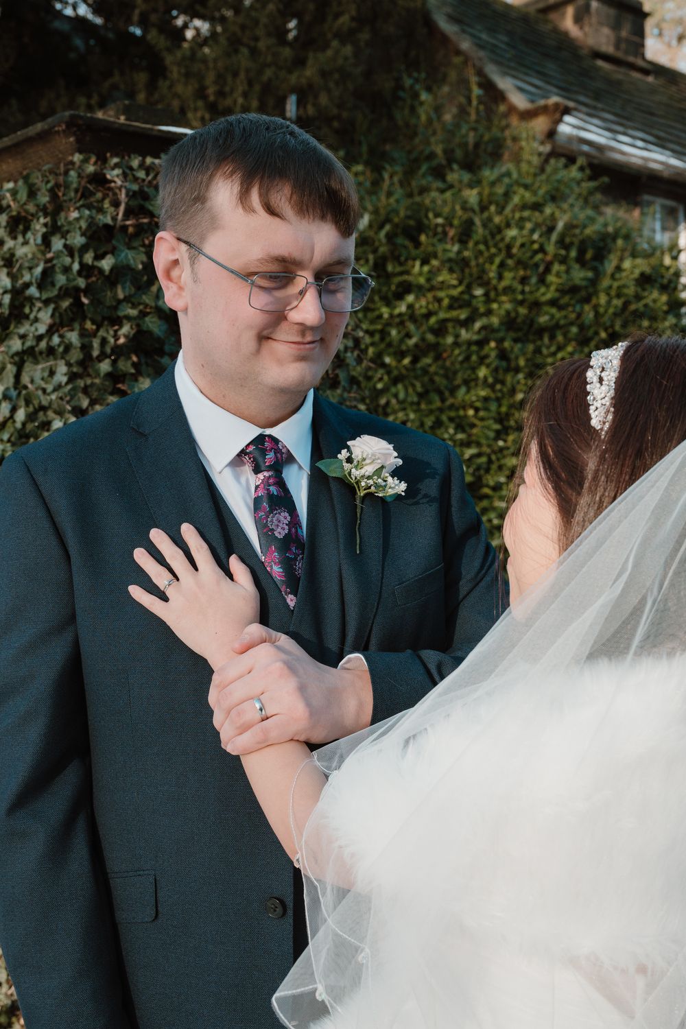 A groom in a dark suit with floral tie and boutonniere smiles at his bride in a veil during their wedding day.