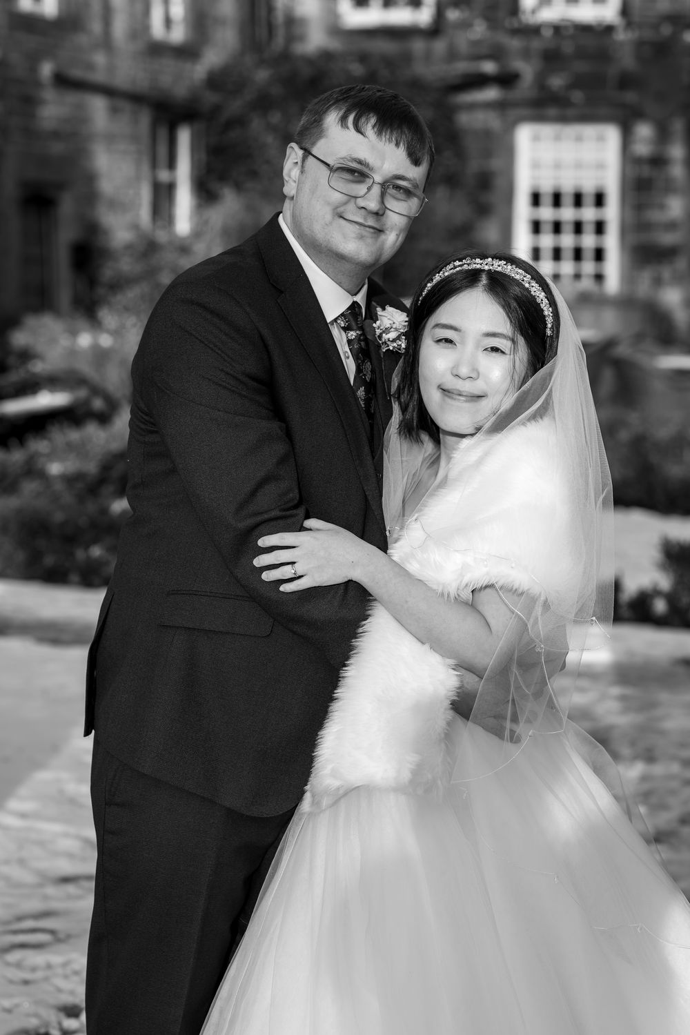 Black and white wedding portrait of a smiling couple, bride in veil and gown, groom in suit, posing outdoors.