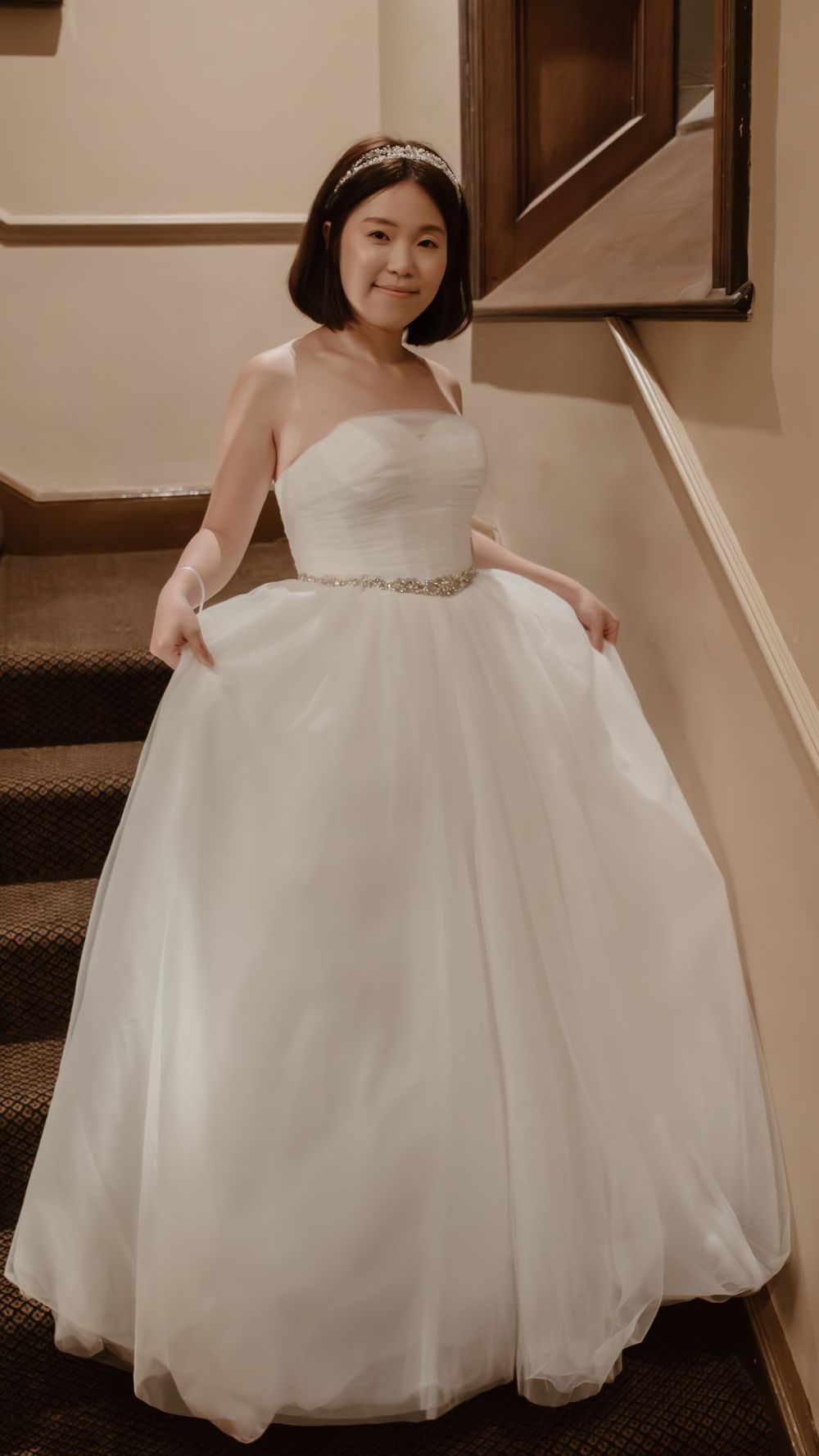 Woman in strapless white ball gown wedding dress with crystal belt and tiara posing on staircase.