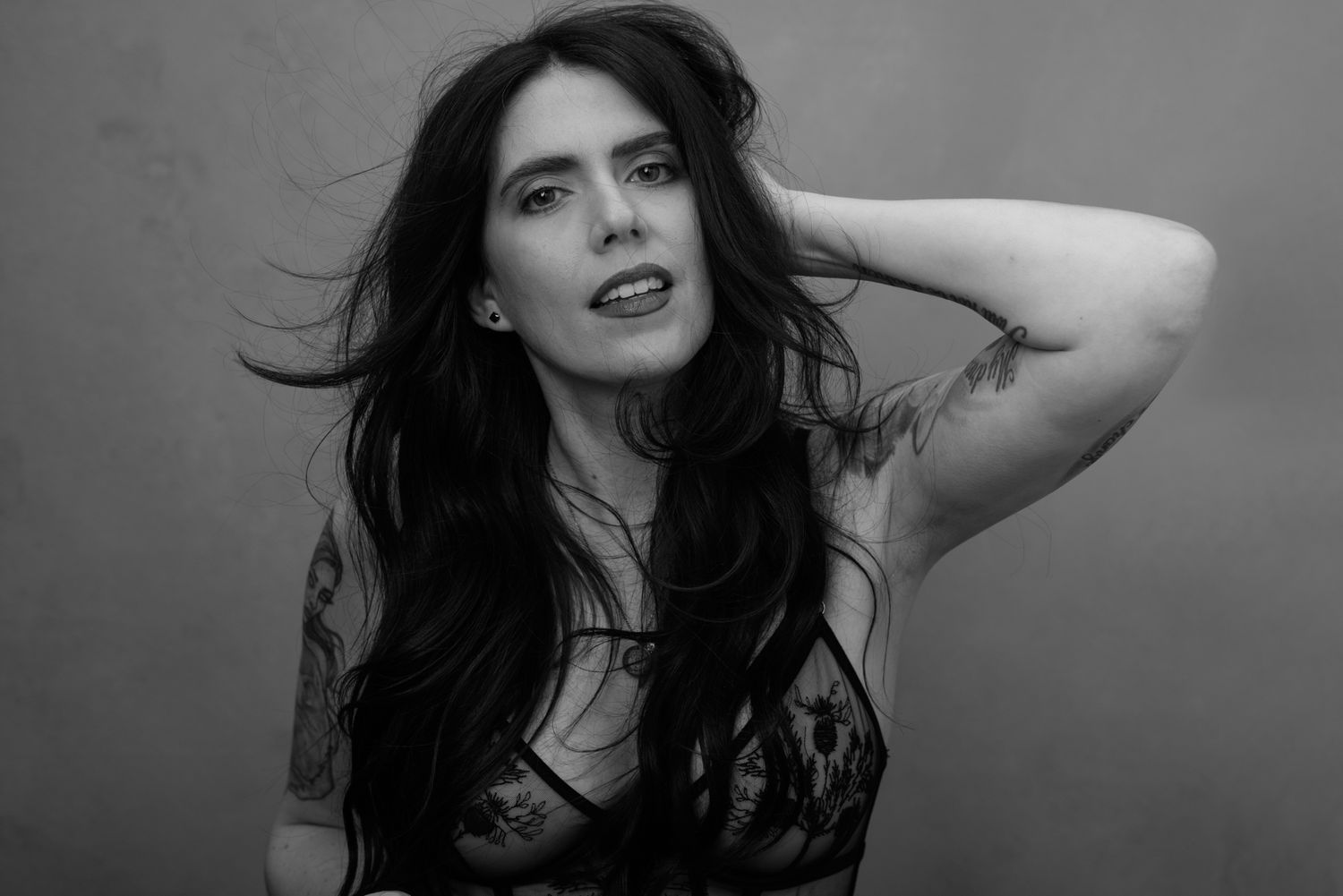 Black and white portrait of a woman with long dark hair, tattoos, posing with hand behind head.