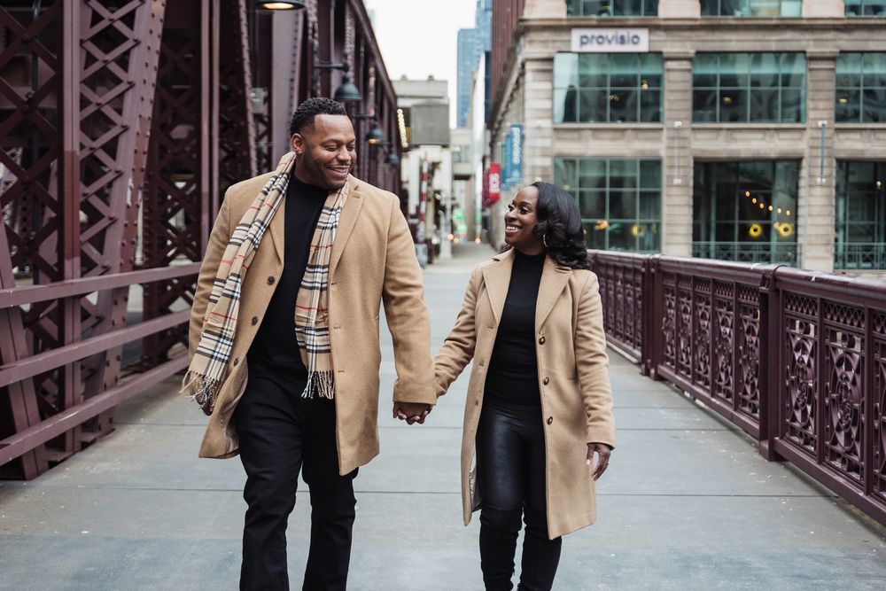 A happy Black couple walks hand-in-hand across a city bridge, both wearing matching beige coats on a cloudy day.