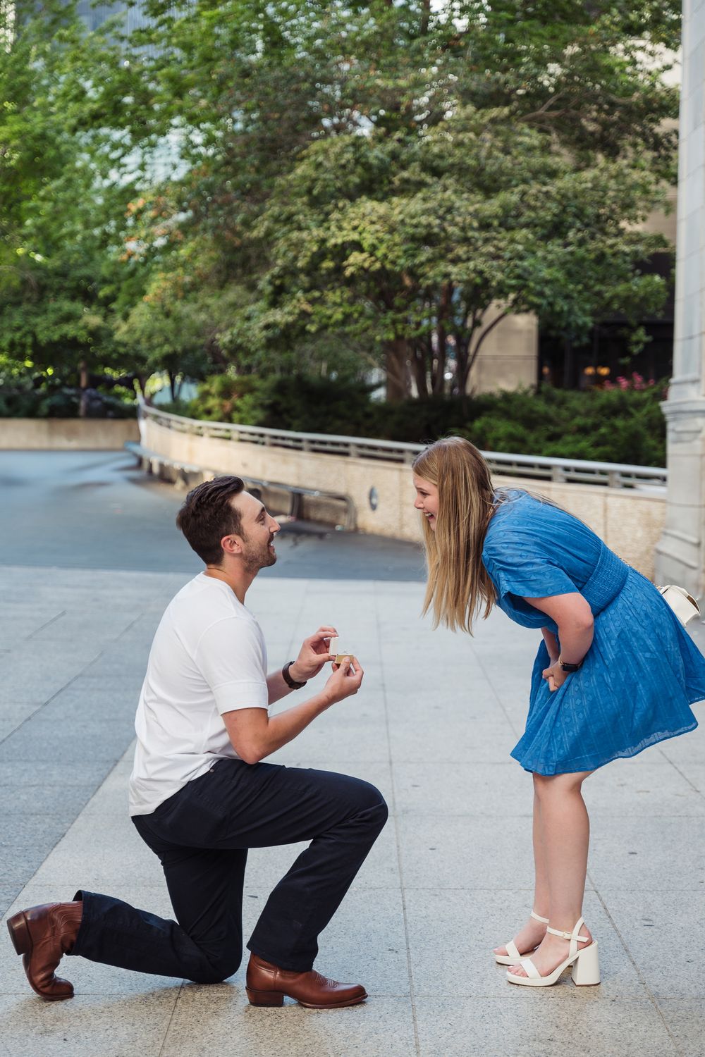 Man kneeling on one knee proposing to woman in blue dress outdoors, romantic marriage proposal moment.