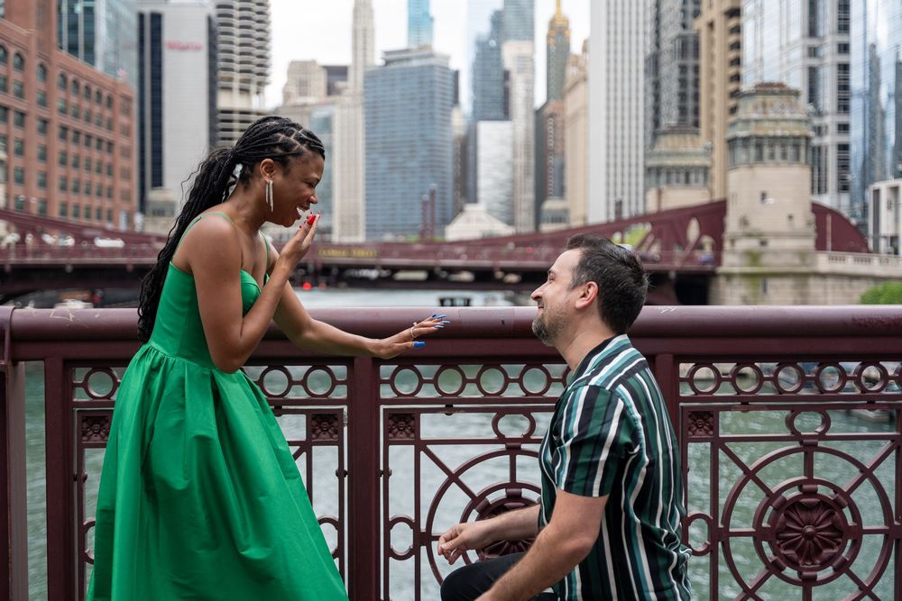Man proposes to woman on bridge over Chicago River, she reacts with surprise, city skyline in background.
