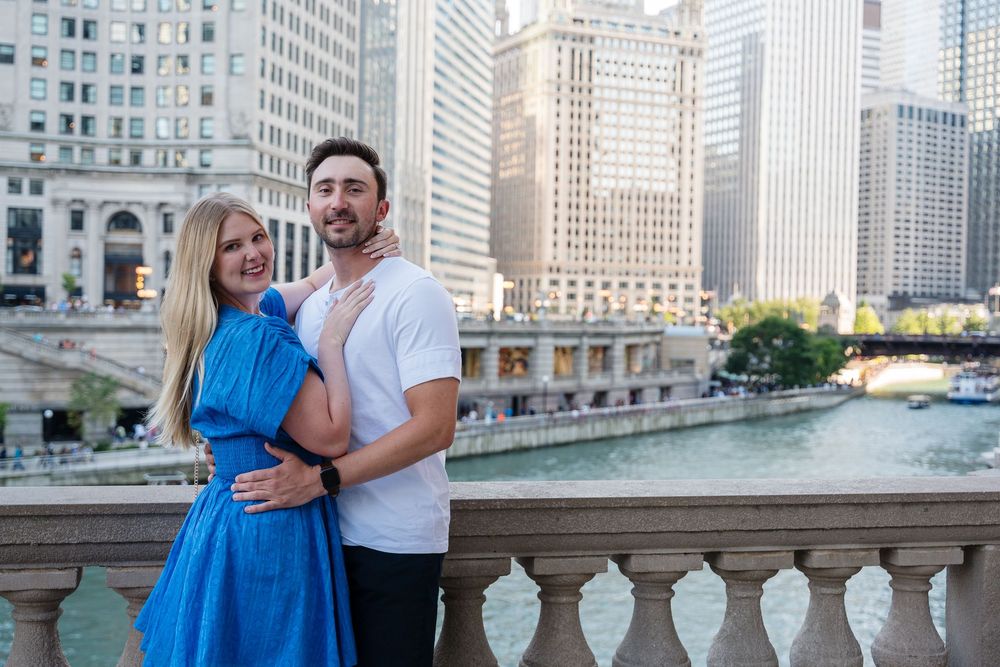 Couple embracing on a bridge overlooking the Chicago River with city skyscrapers in the background.