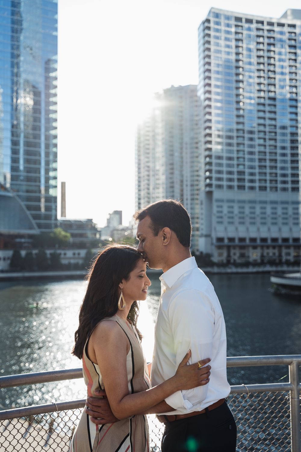 Romantic couple embracing on a city bridge with modern skyscrapers and a river in the background on a sunny day.