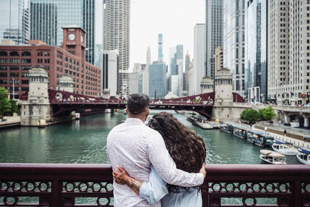 Couple embracing on a bridge overlooking the Chicago River with city skyline and red bridge in background.
