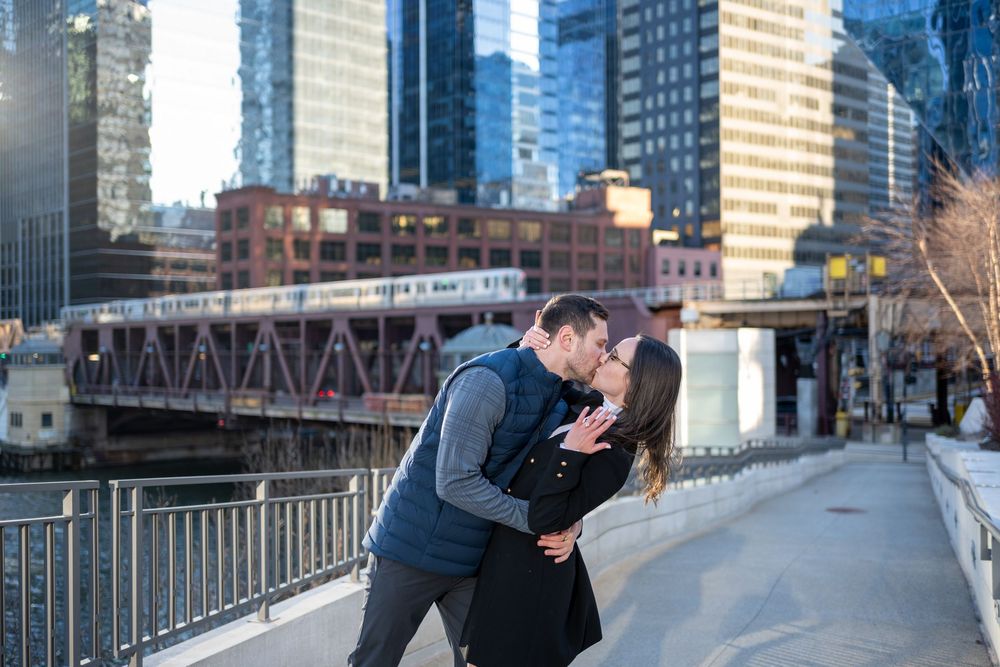 Couple sharing a romantic dip kiss on a Chicago riverwalk bridge with city skyline and elevated train in background.