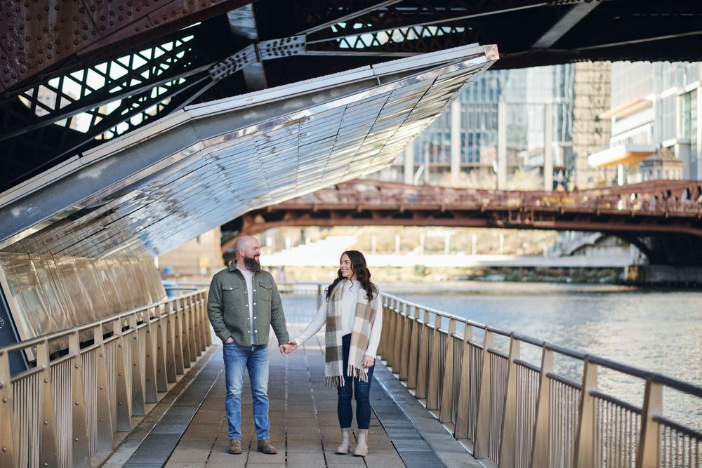 Couple holding hands walking on a riverwalk bridge in Chicago with urban architecture and waterway in the background.