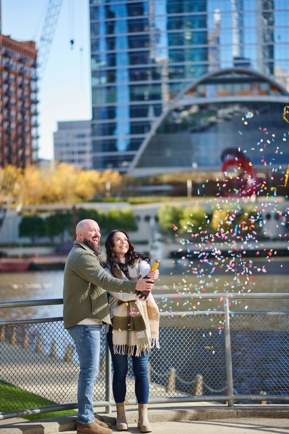 Happy couple celebrating with confetti by a urban waterfront, wearing cozy fall outfits with city skyline backdrop.