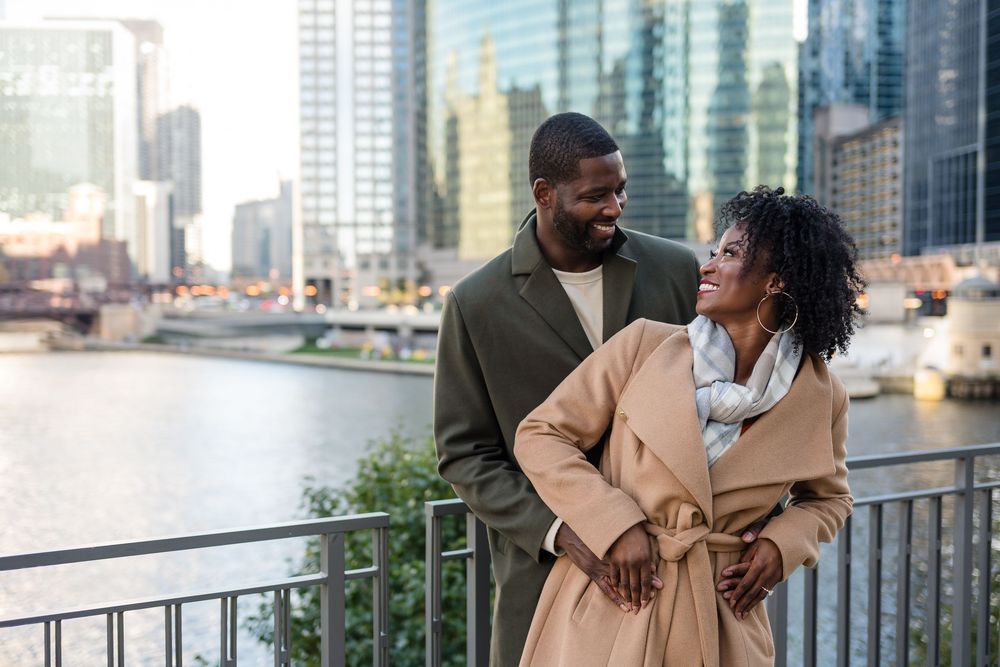 A happy Black couple smiles and embraces on a city riverwalk with modern skyscrapers in the background.