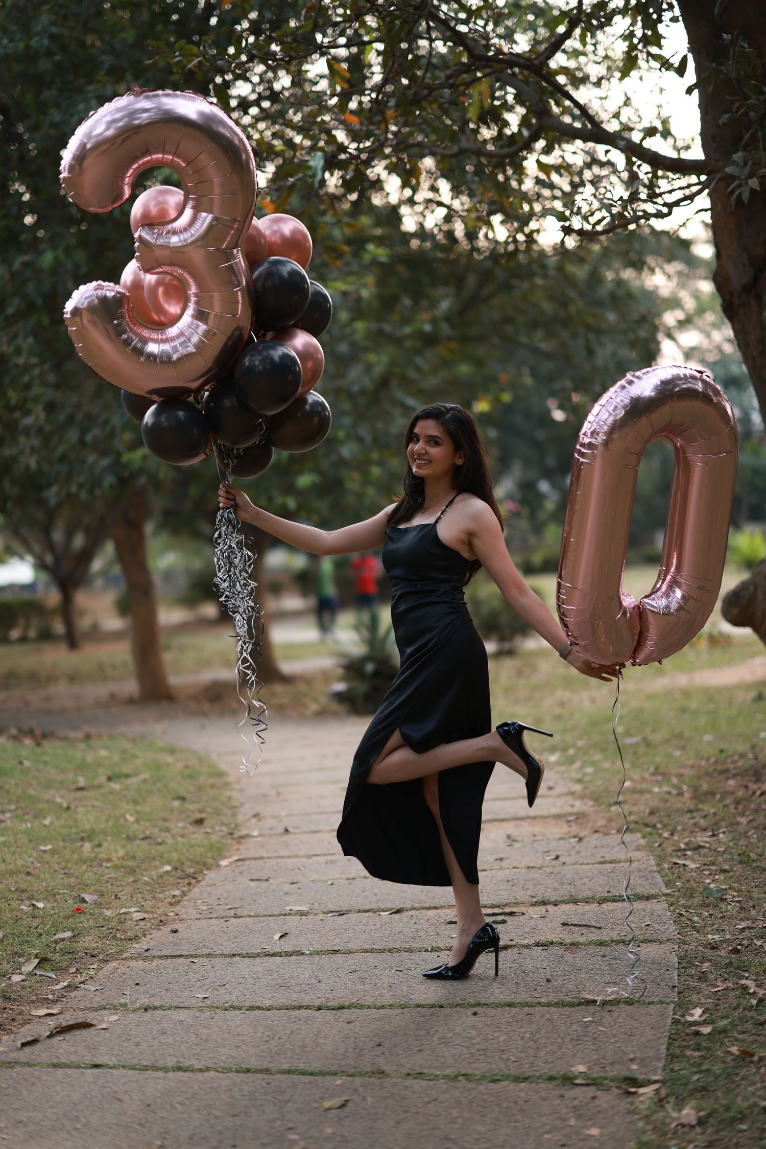 Woman in black dress celebrating 30th birthday in park, holding rose gold and black number 30 balloons.