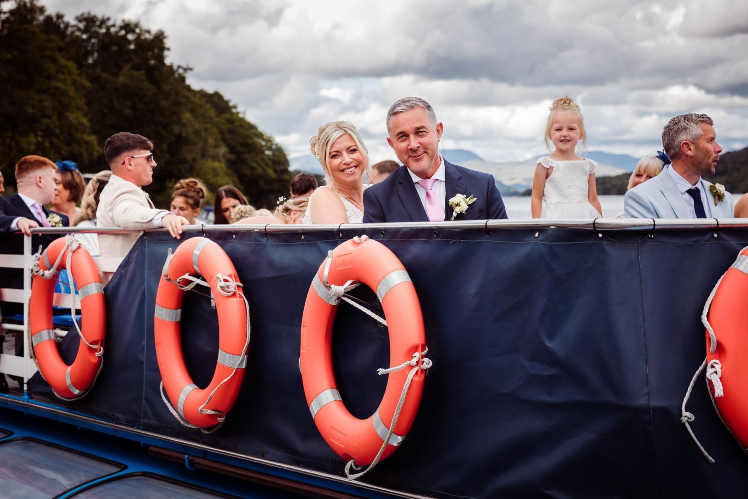 Wedding couple smiling on a boat with orange life rings, surrounded by guests on a scenic lake.