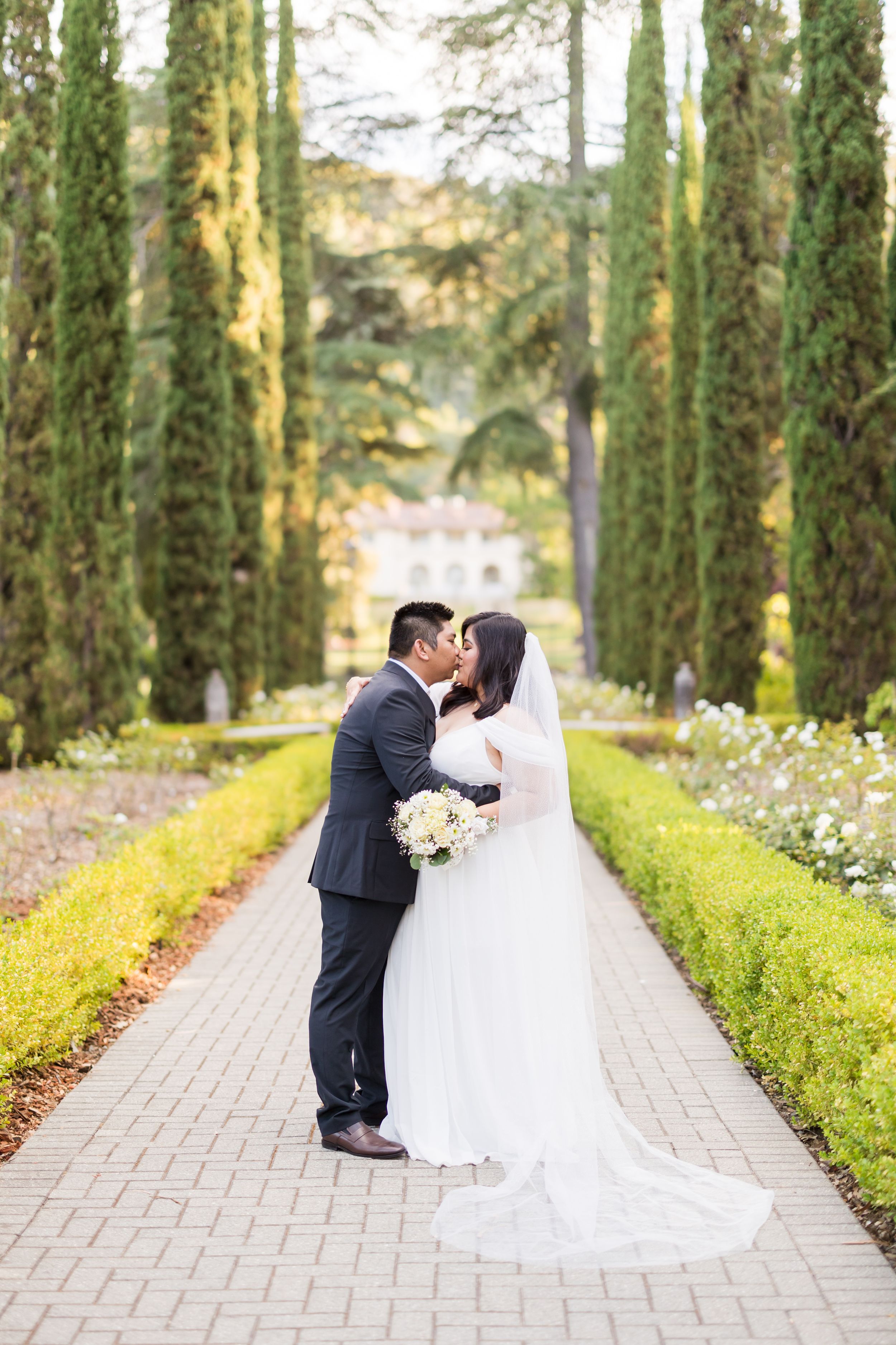 Bride and groom kissing on a garden pathway lined with tall cypress trees and manicured hedges at an elegant wedding venue.