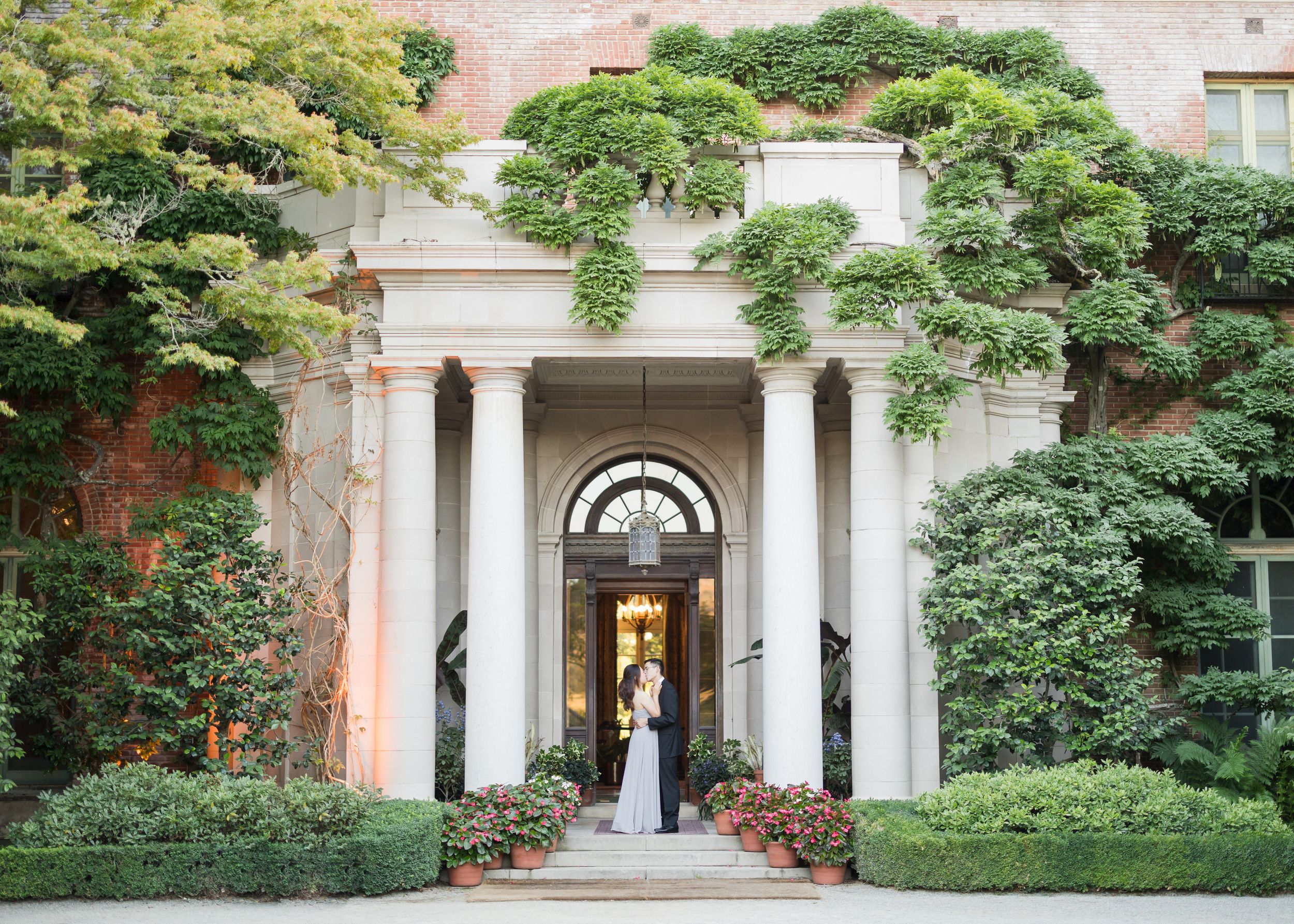 Couple embracing at elegant mansion entrance with white columns, lush greenery, and brick facade for a romantic wedding photo.