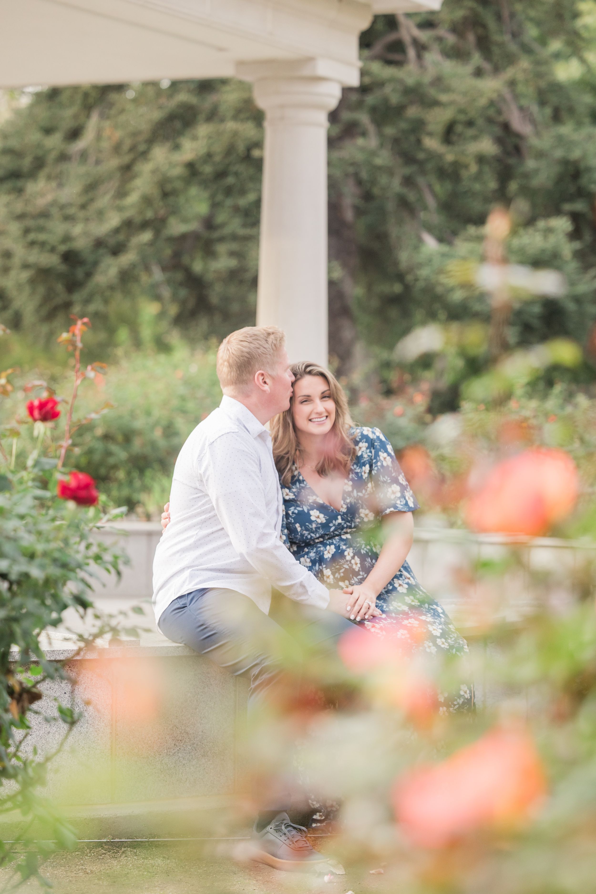 Couple sitting together among blooming roses by white columns in a lush garden, smiling warmly at each other.