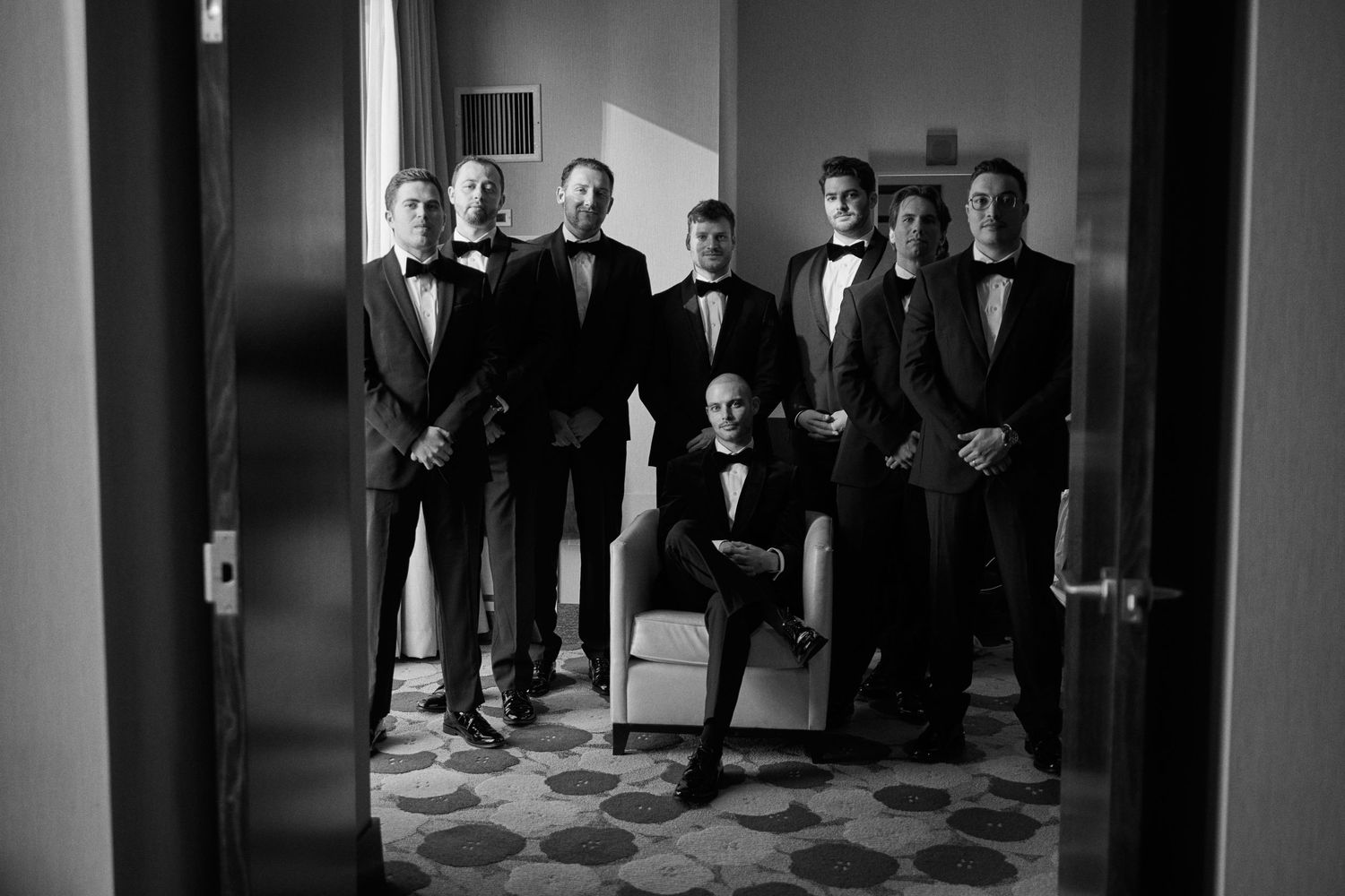 Black and white photo of six men in tuxedos posing formally in a doorway for a wedding groomsmen portrait.
