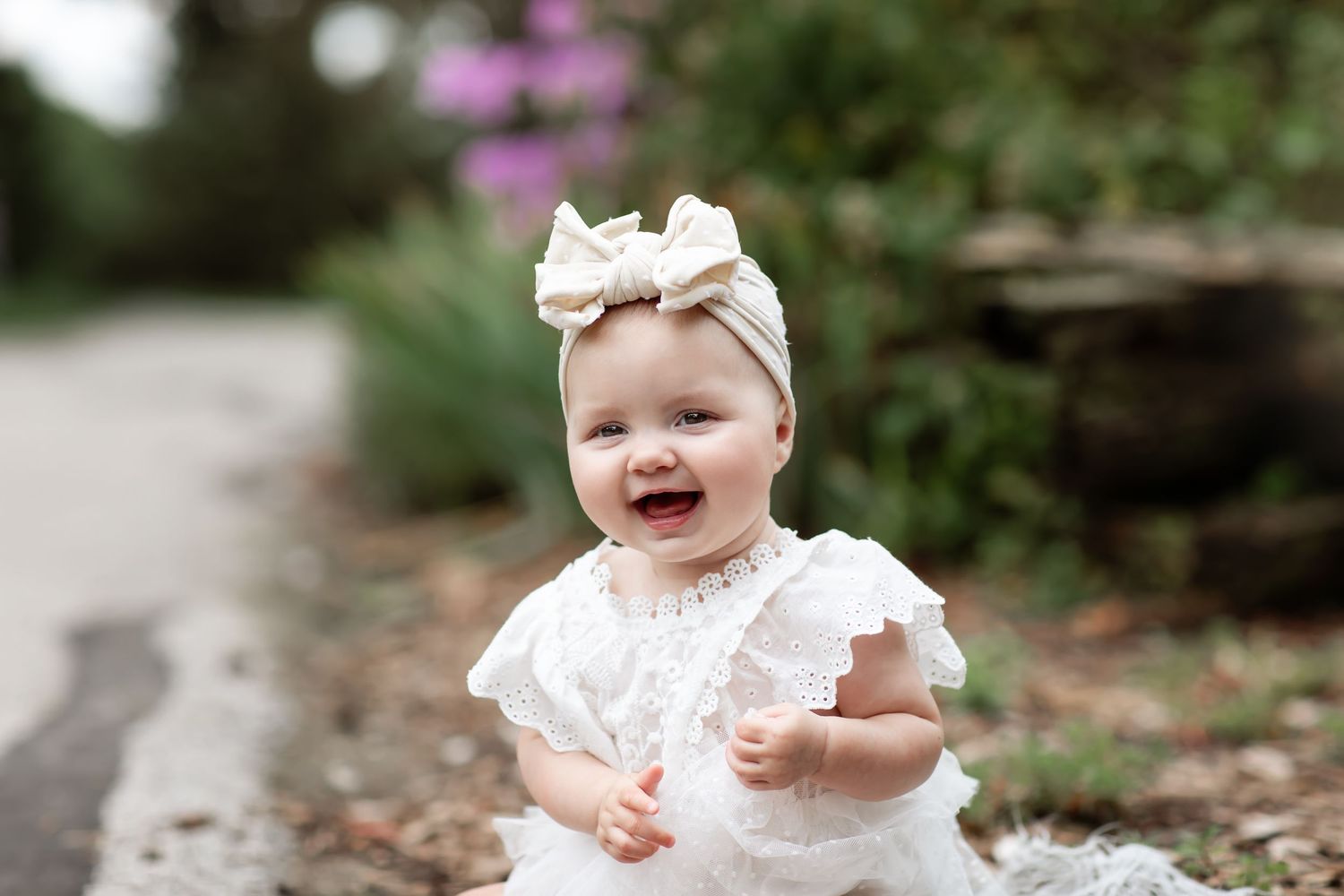 Adorable baby girl in white lace dress and bow headband sitting outdoors in a lush garden setting.