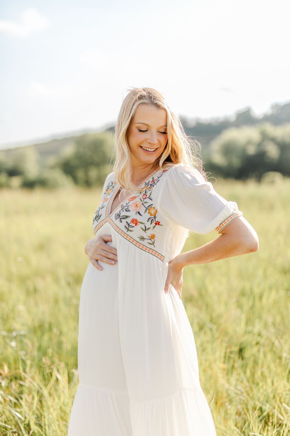Glowing pregnant woman in a floral white dress smiling down at her baby bump outdoors.