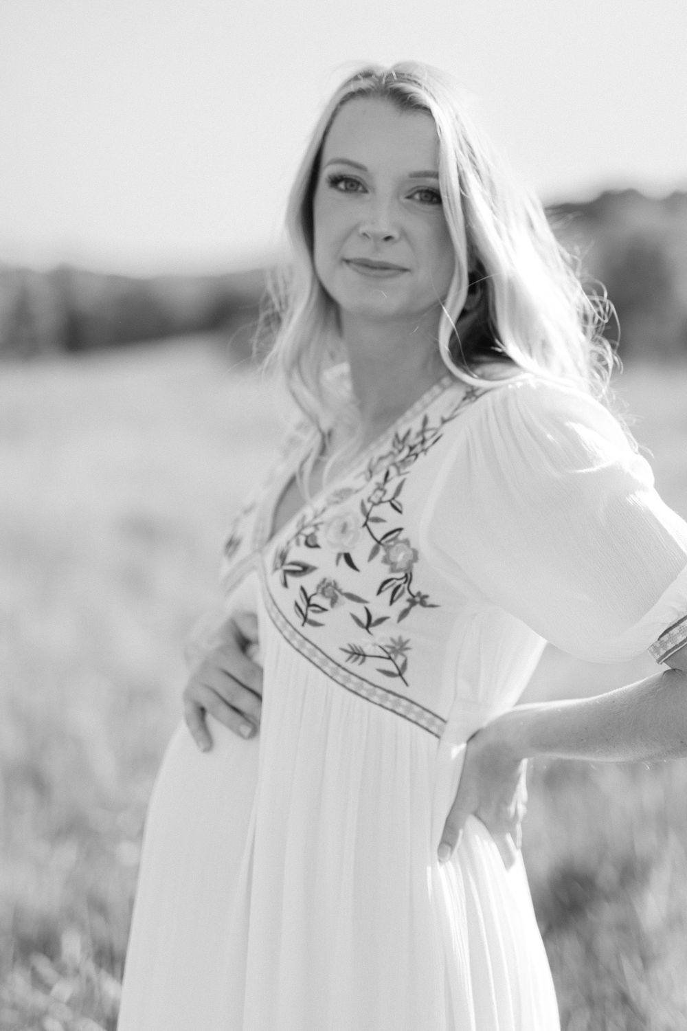 Black and white portrait of a pregnant woman in a floral embroidered white dress in a field.