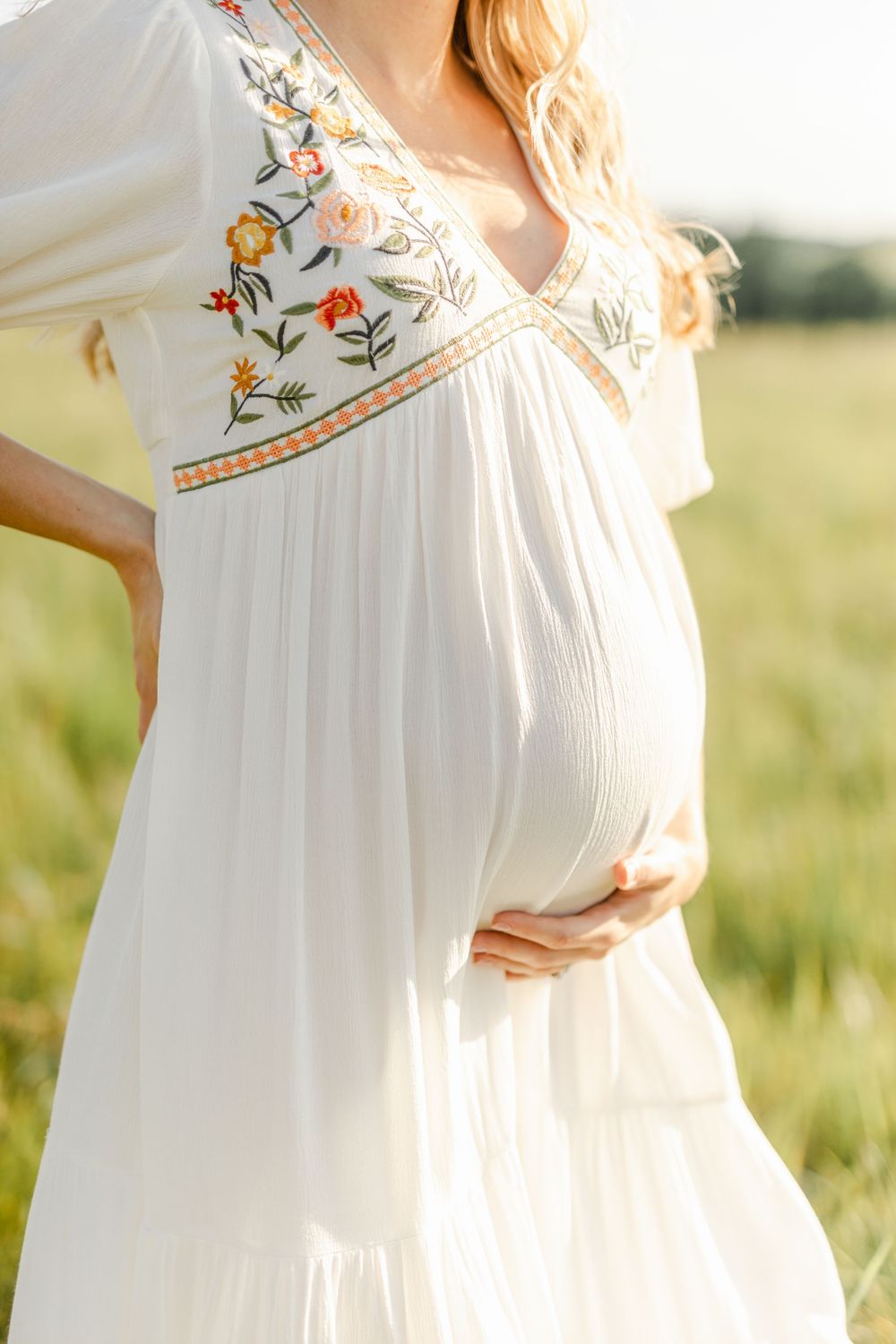 Close-up of a pregnant woman cradling her baby bump in a white floral embroidered dress outdoors.