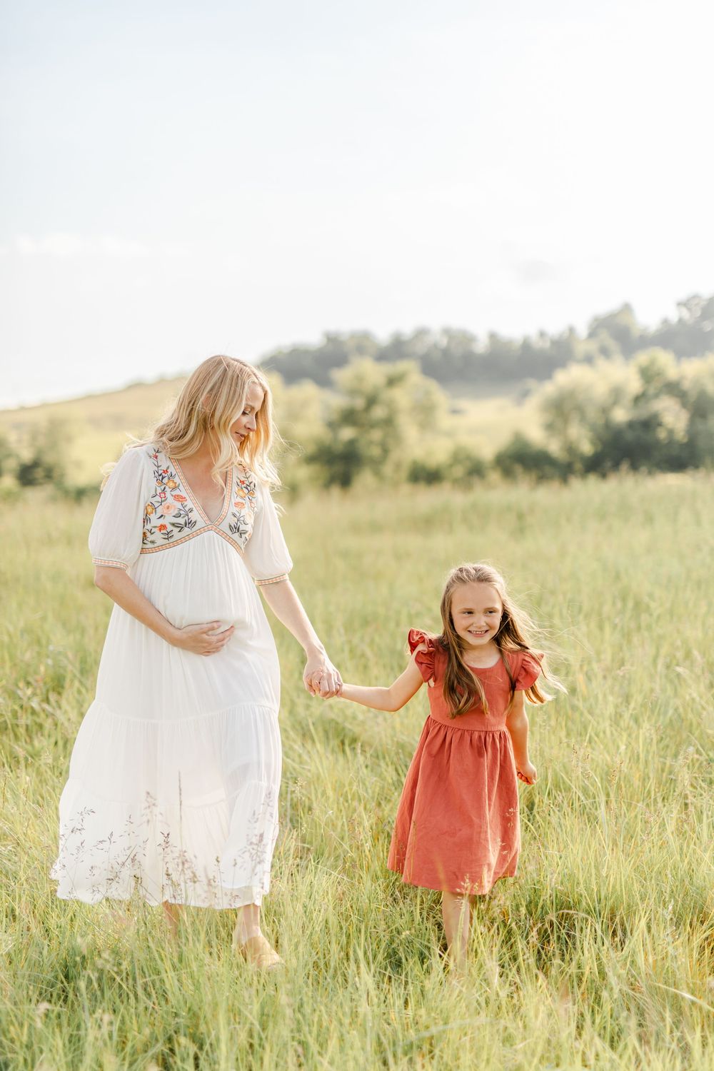 Pregnant woman in white dress holding hands with a young girl in a rust dress in a green field.