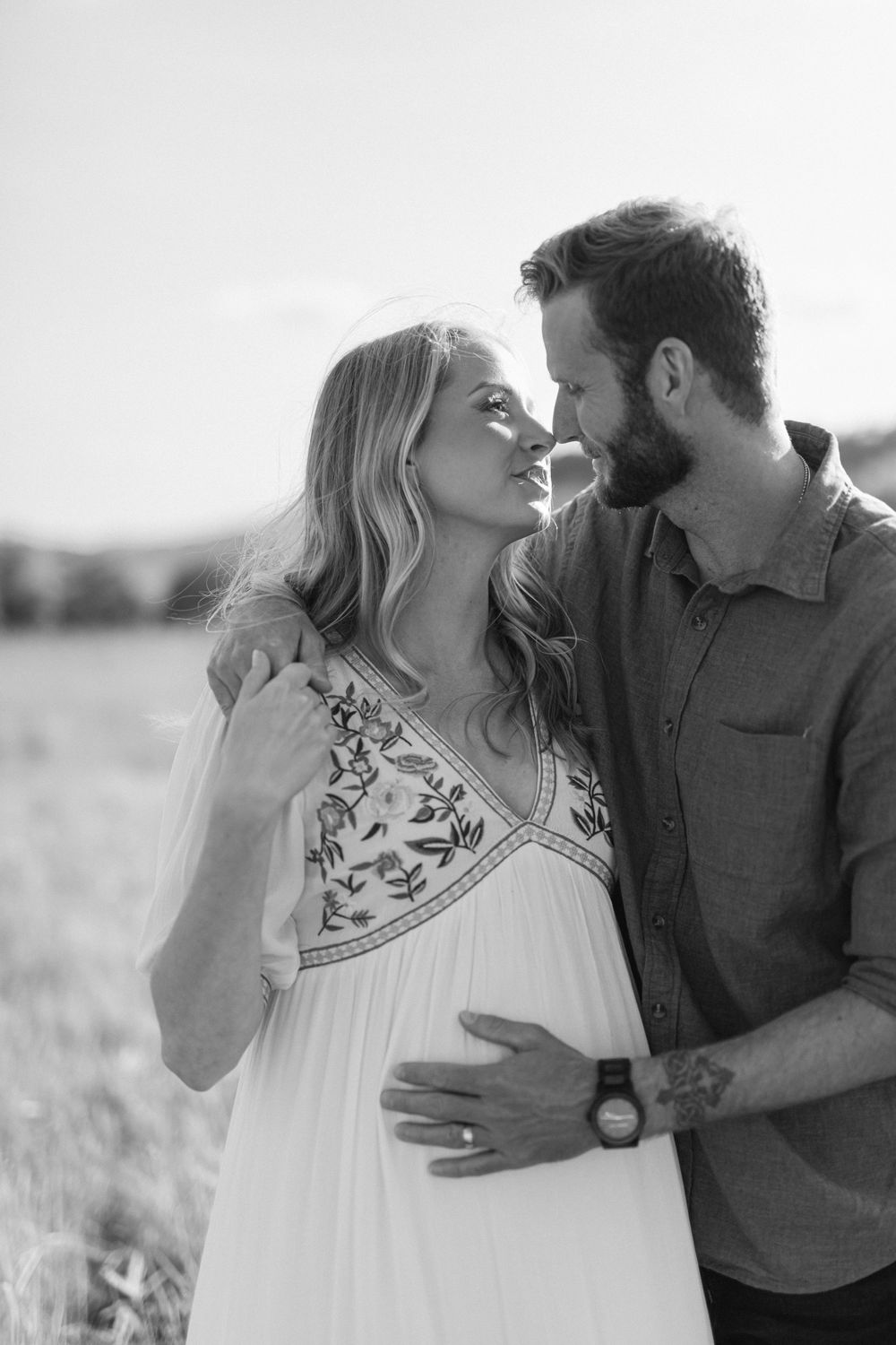 Black and white maternity photo of couple embracing in field, man holding pregnant womans belly.